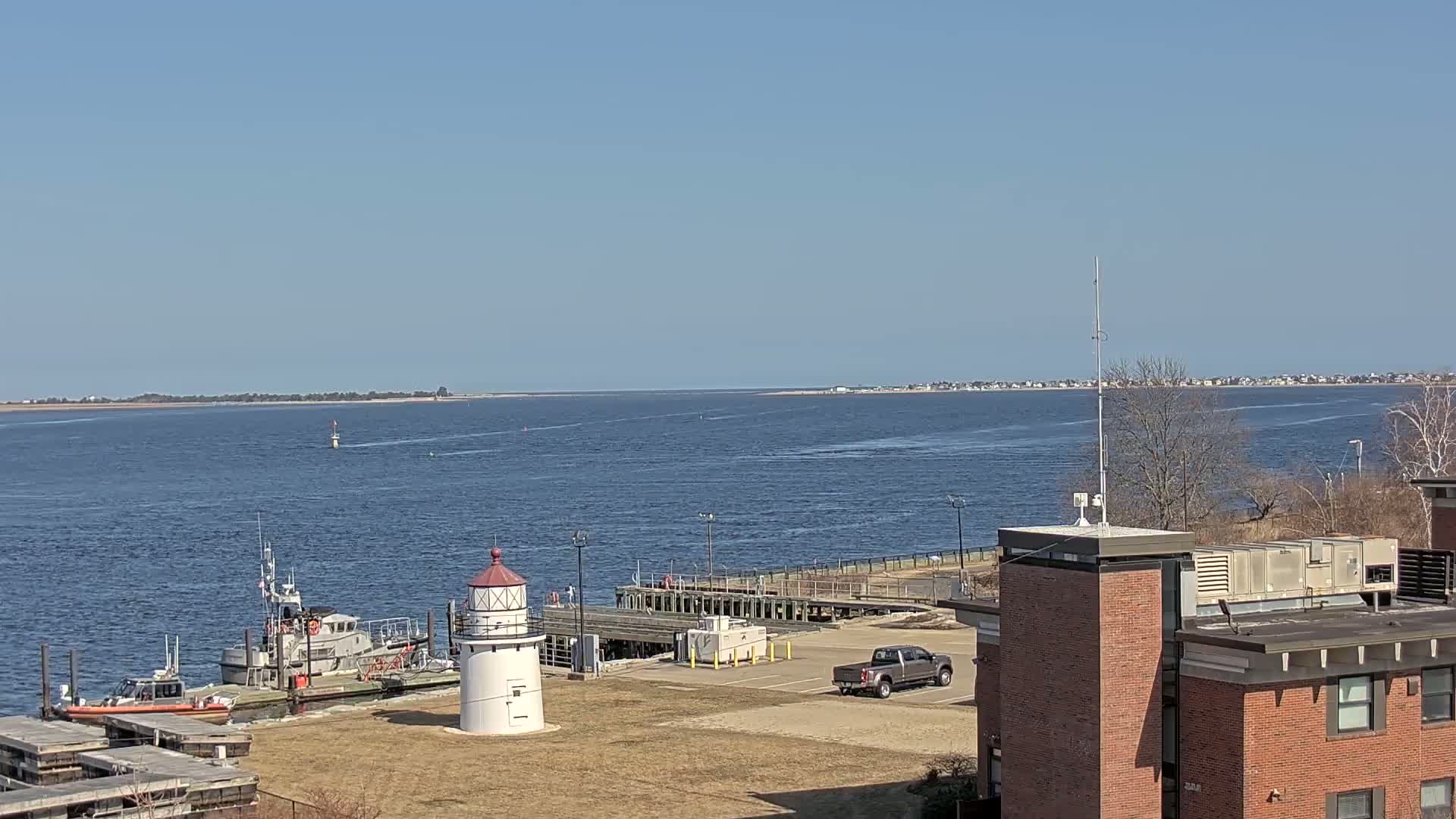 A harbor scene on a sunny day shows a small lighthouse, two Coast Guard boats, a parking lot with a truck, and a brick building, all overlooking a calm body of water with a distant shoreline.