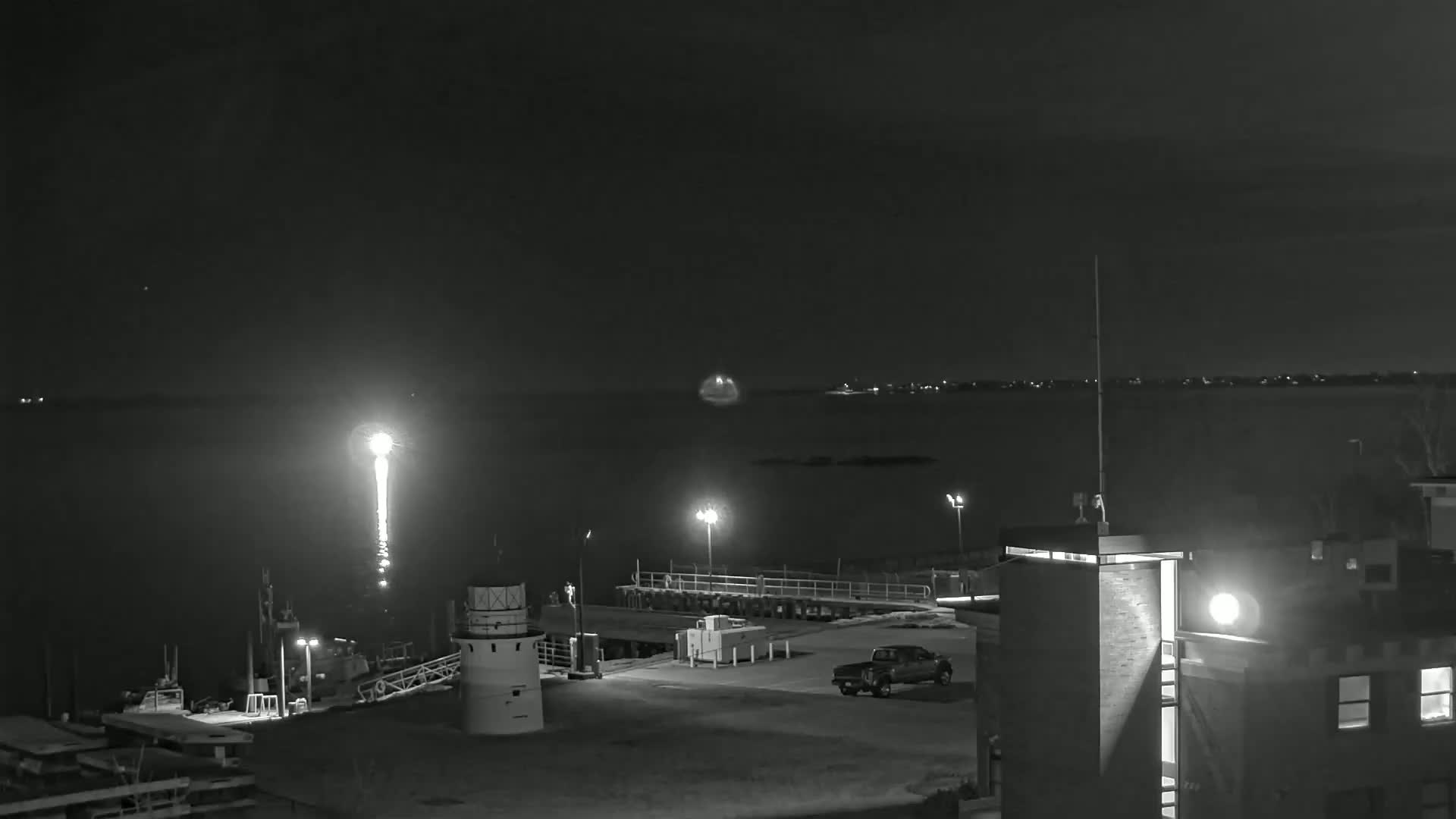 A nighttime, black and white view shows a small harbor with a lighthouse, a few boats, a pickup truck, and a building under a clear, dark sky.