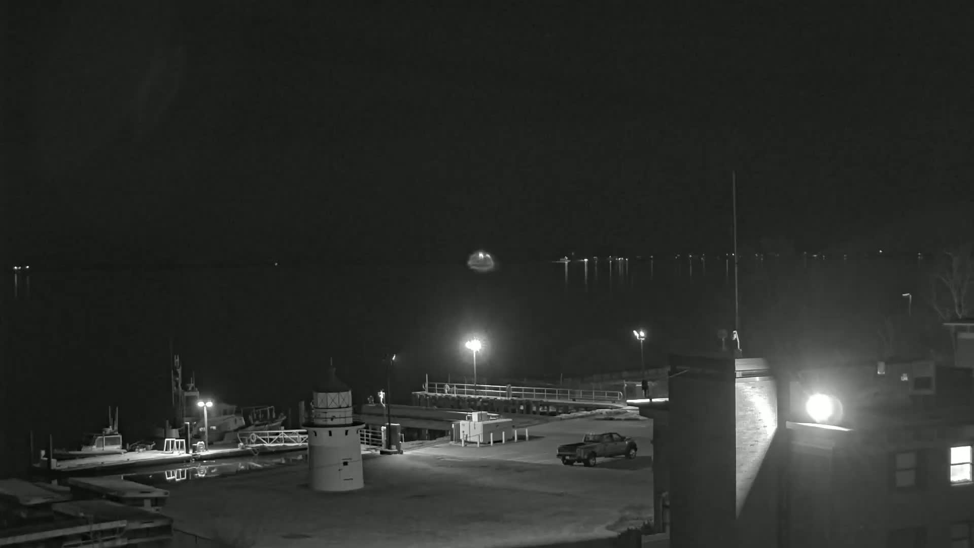A nighttime, black-and-white view of a harbor with a small lighthouse, docked boats, and a pickup truck parked in a lot near a building, all illuminated by artificial light.