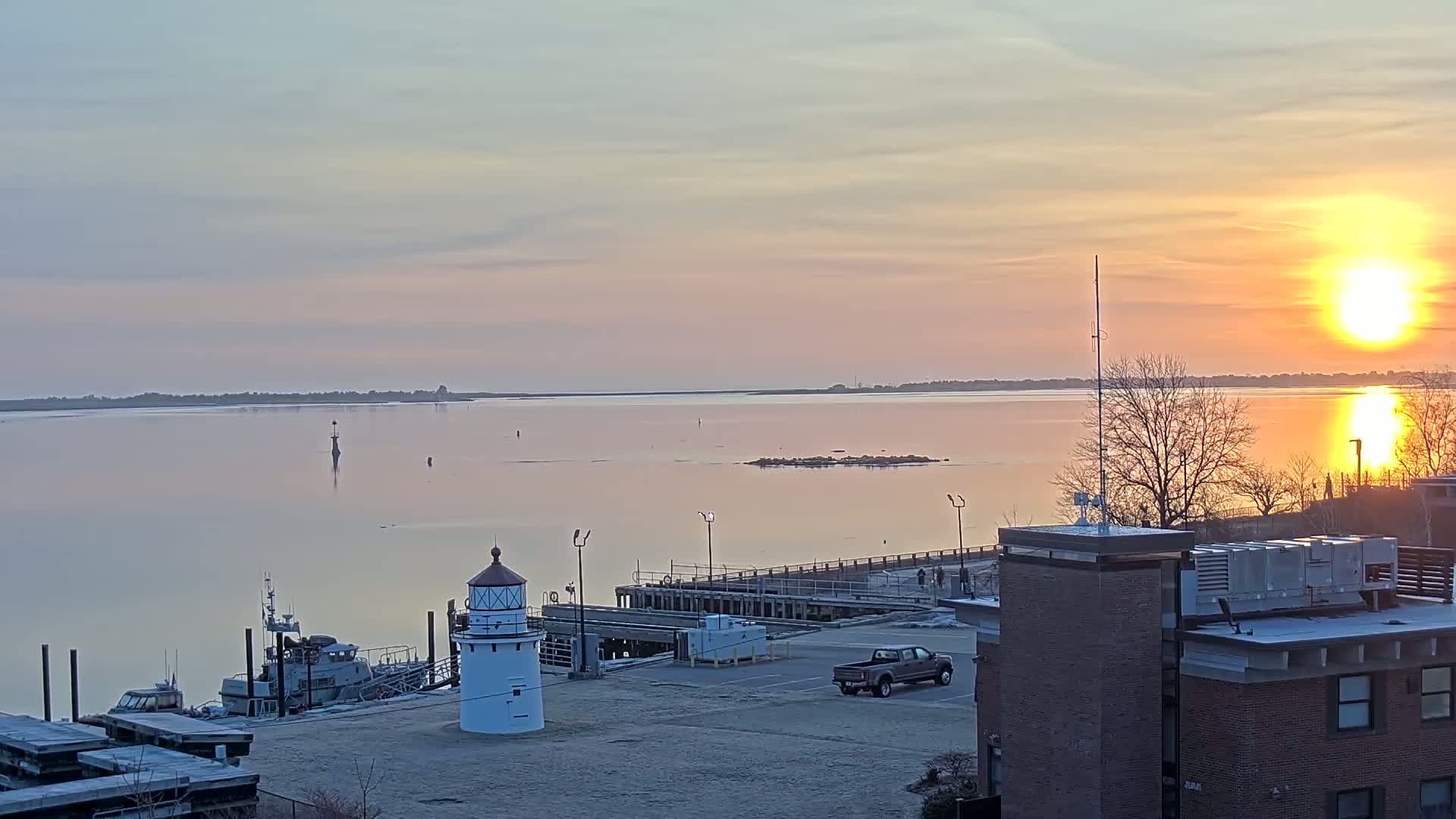 A calm body of water reflects a setting sun, with a small lighthouse, boats, and a building visible on a dock in the foreground.