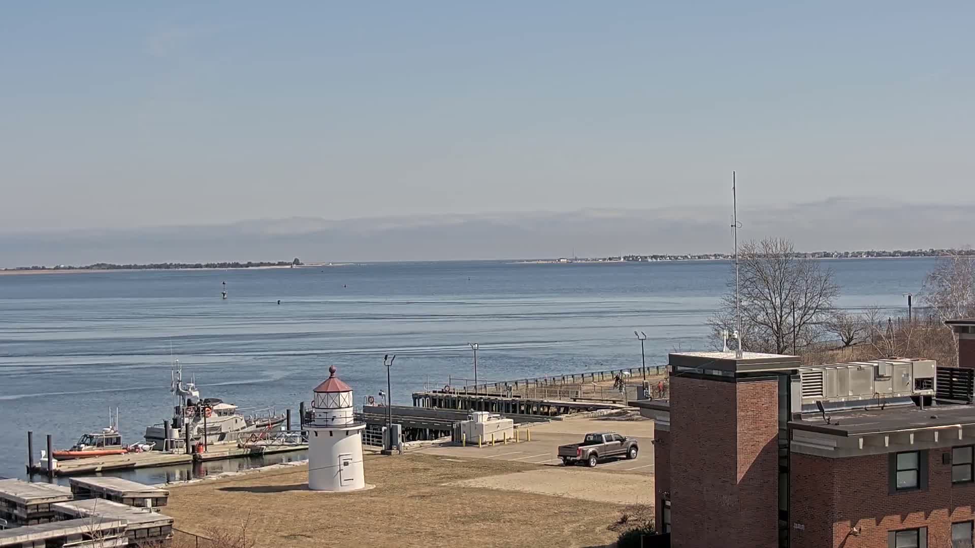 A white lighthouse stands near a harbor with a Coast Guard boat docked, under a clear blue sky and calm water, with a distant shoreline visible.