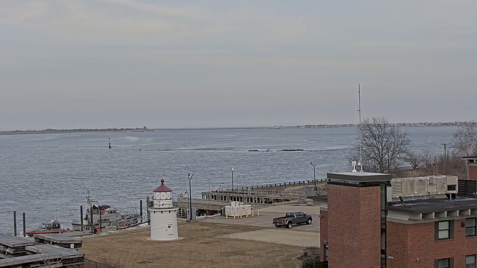 A small lighthouse stands near a harbor with a Coast Guard boat, a pickup truck in a parking lot, and a brick building overlooking a calm, gray body of water under an overcast sky.