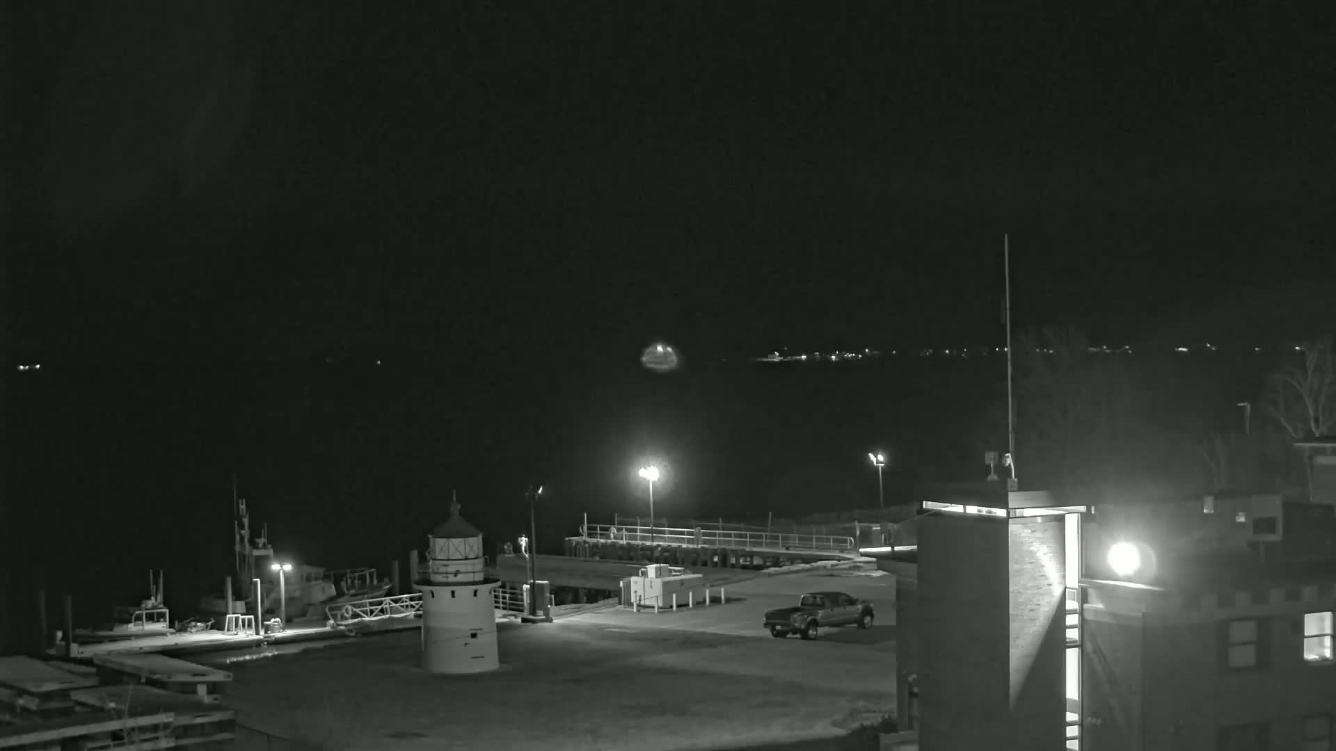 A nighttime, black-and-white view of a harbor with a lighthouse, docked boats, a truck in a parking lot, and a building under a dark sky.