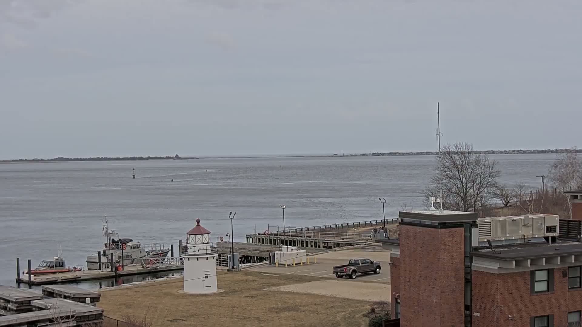 A small lighthouse and Coast Guard boats are docked at a harbor on an overcast day, with a wide expanse of calm water and distant shoreline visible in the background.