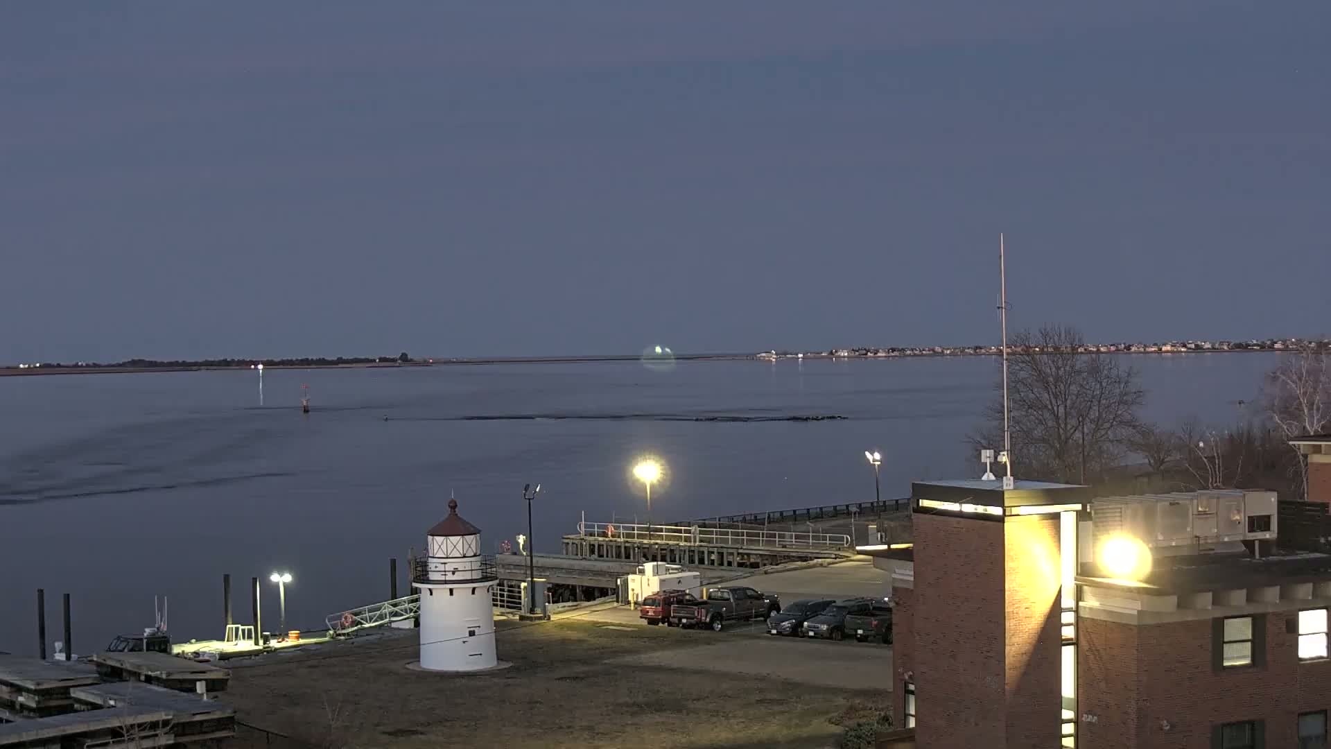 A white lighthouse sits on a pier next to a body of water under a twilight sky, with a brick building and parked cars in the foreground.