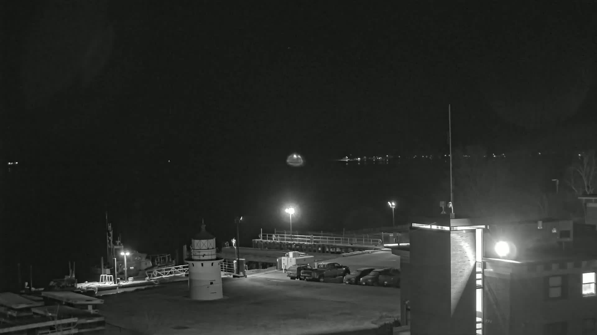 A nighttime view of a harbor with a lighthouse, several docked boats, a parking lot with cars, and a building, under a dark, clear sky.