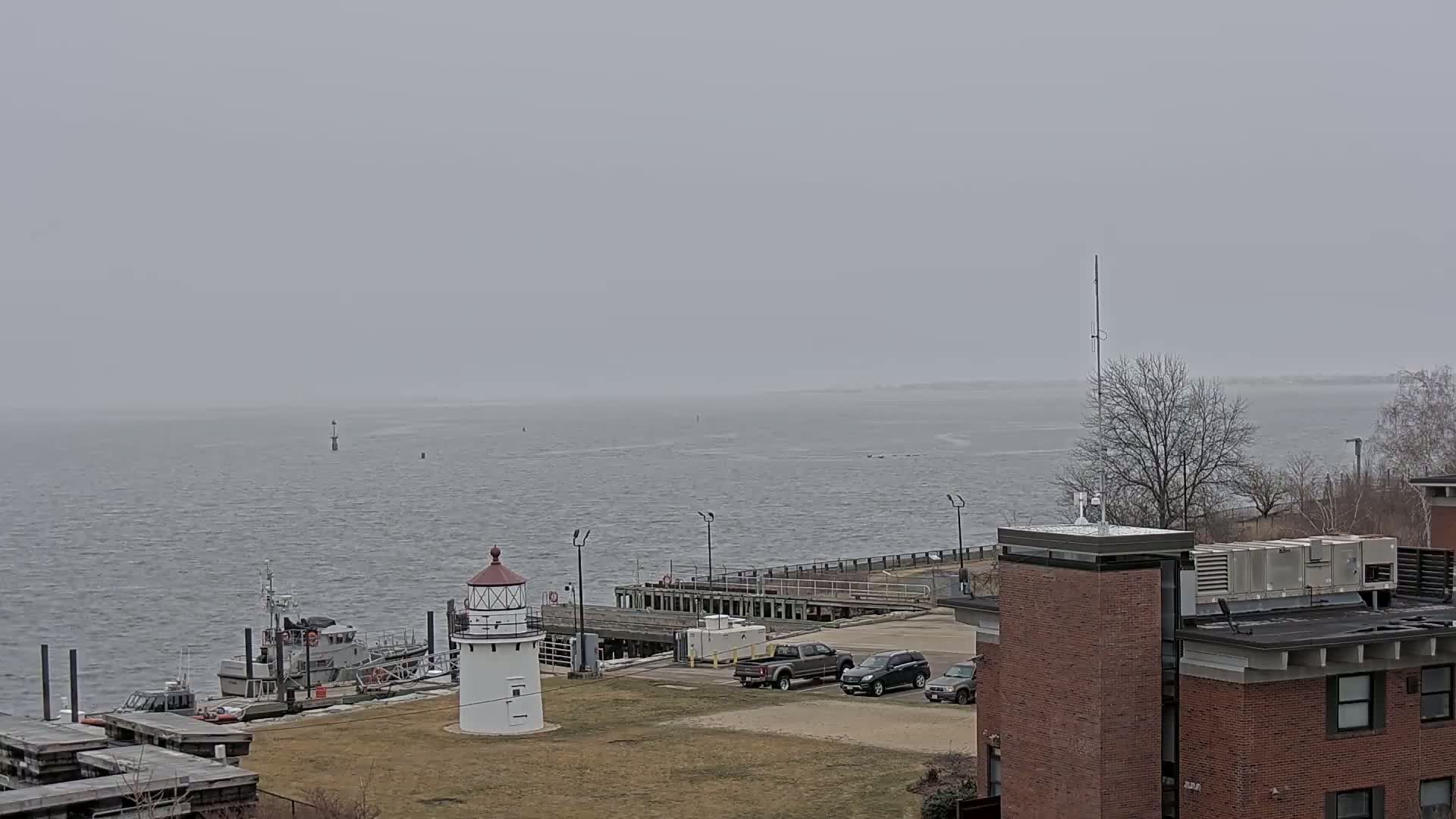 A white lighthouse stands near a dock with several small boats and cars in a parking lot, all overlooking a gray, choppy body of water under an overcast sky.