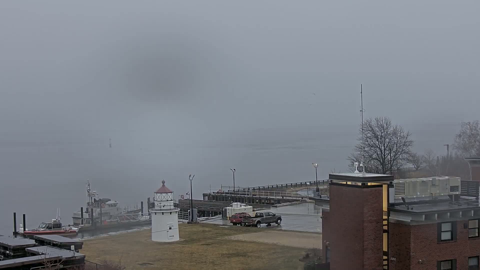 A small lighthouse and docked boats are visible on a foggy day next to a brick building.