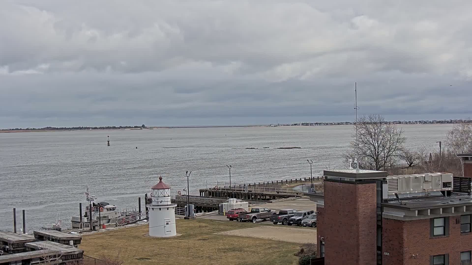 A small lighthouse stands near a dock with several vehicles parked nearby, overlooking a calm body of water under a cloudy sky.