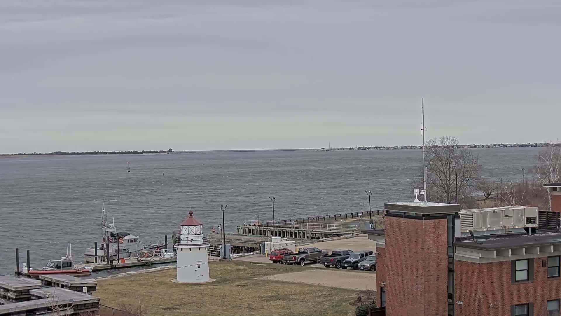 A small lighthouse and several vehicles are situated near a dock with boats on a choppy, grey-watered body of water under an overcast sky.