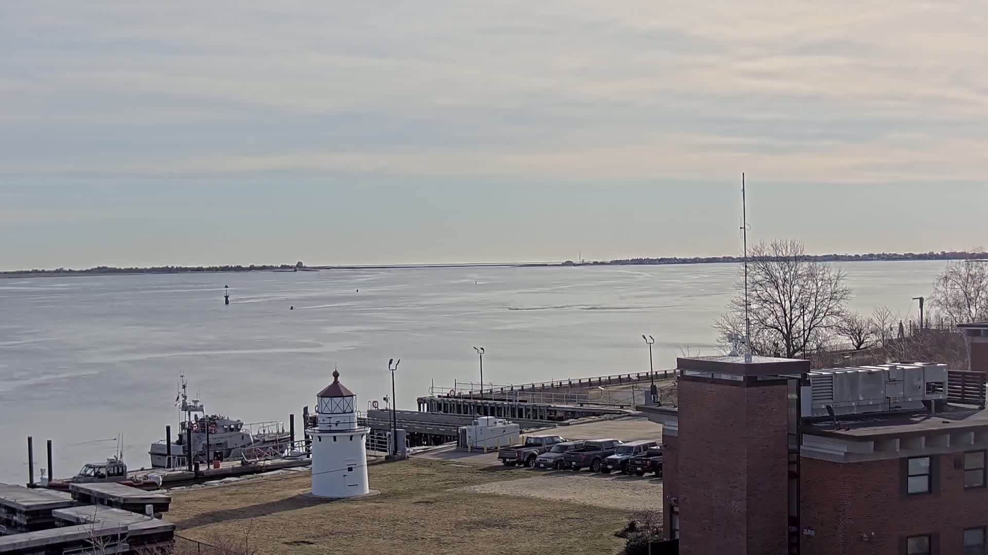 A small white lighthouse stands near a harbor with a docked boat, several parked cars, and a brick building, all overlooking a partially ice-covered bay under a mostly cloudy sky.