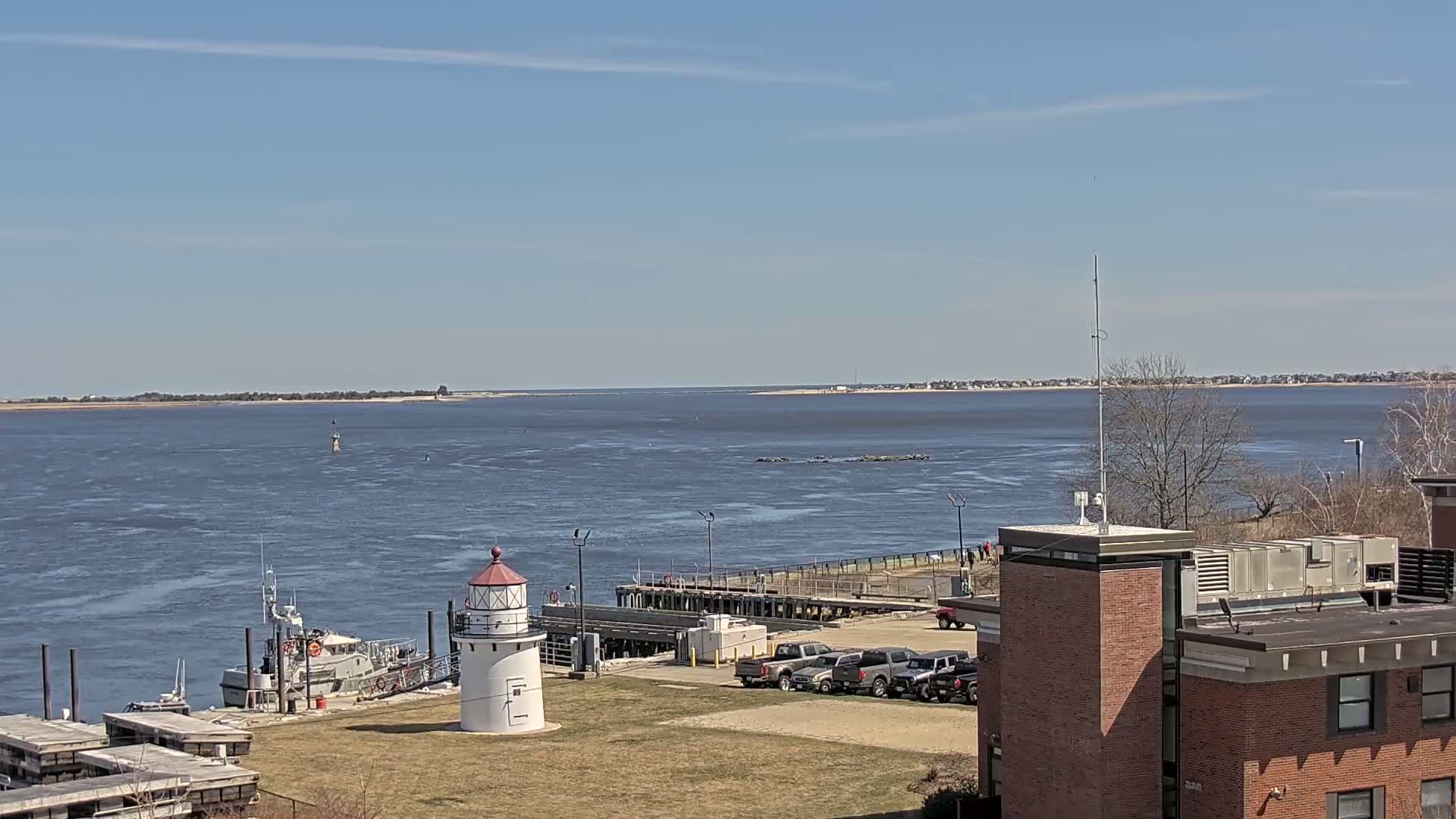 A small lighthouse stands near a docked boat on a calm, sunny day overlooking a wide body of water.