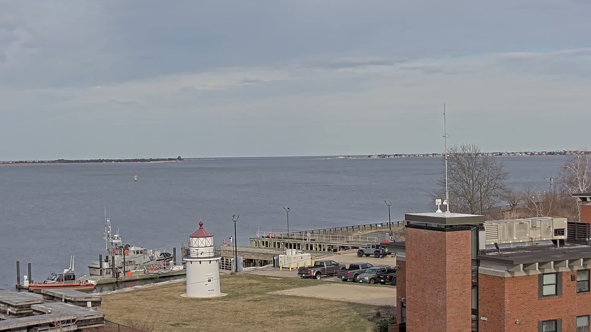 A small lighthouse stands near a dock with several boats and vehicles, overlooking a calm body of water under a cloudy sky.