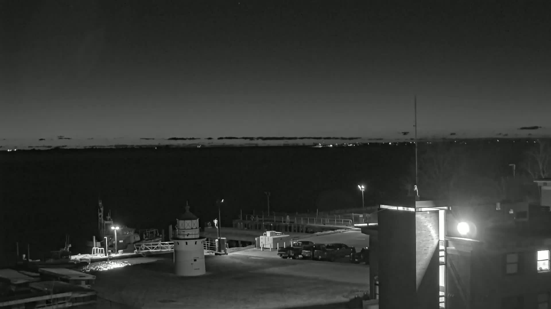 A nighttime, black and white view of a lighthouse and harbor with several vehicles parked nearby, under a mostly clear sky.