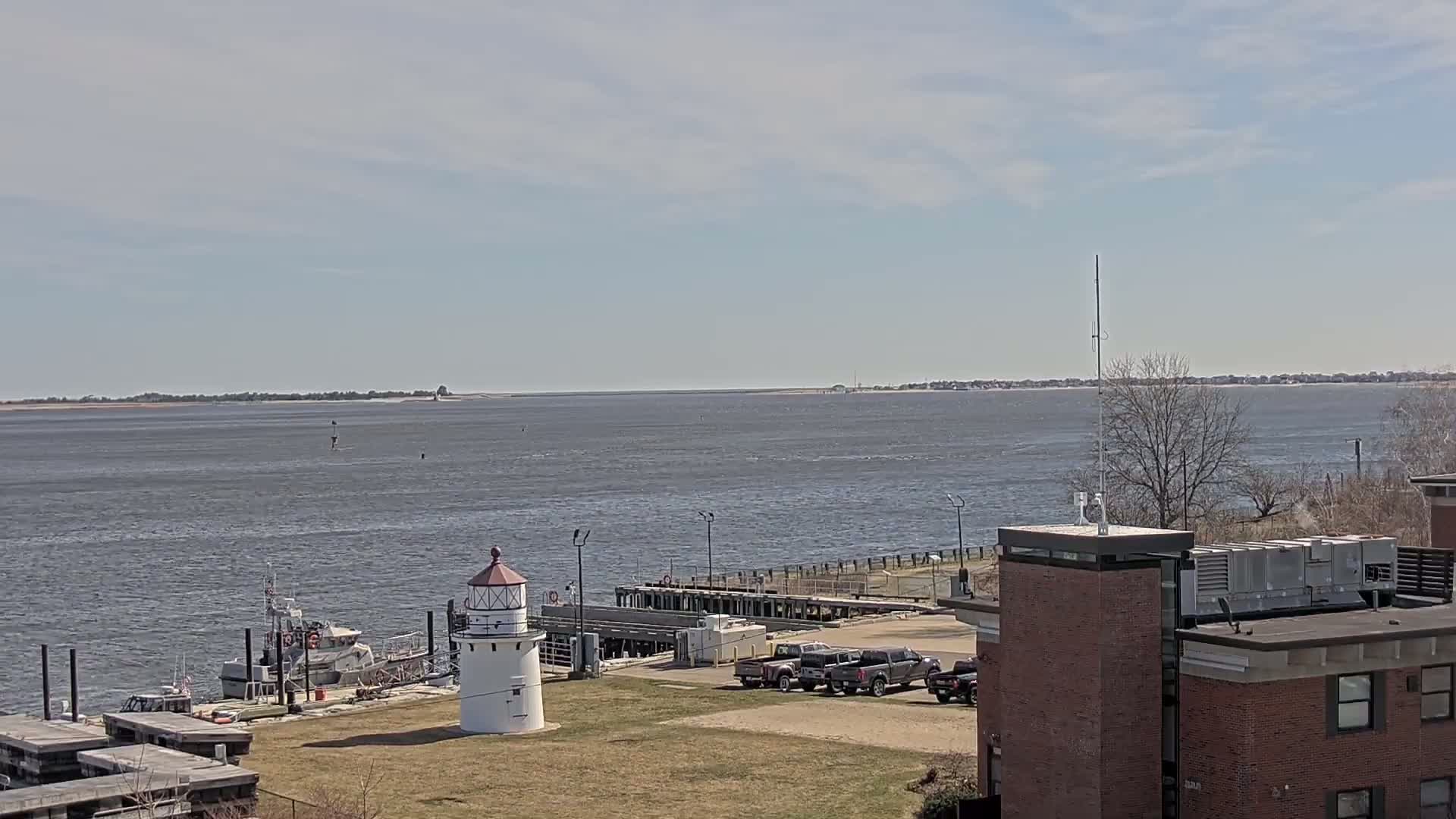 A body of water stretches to a distant shoreline under a partly cloudy sky, with a lighthouse, docked boats, and a building visible in the foreground.