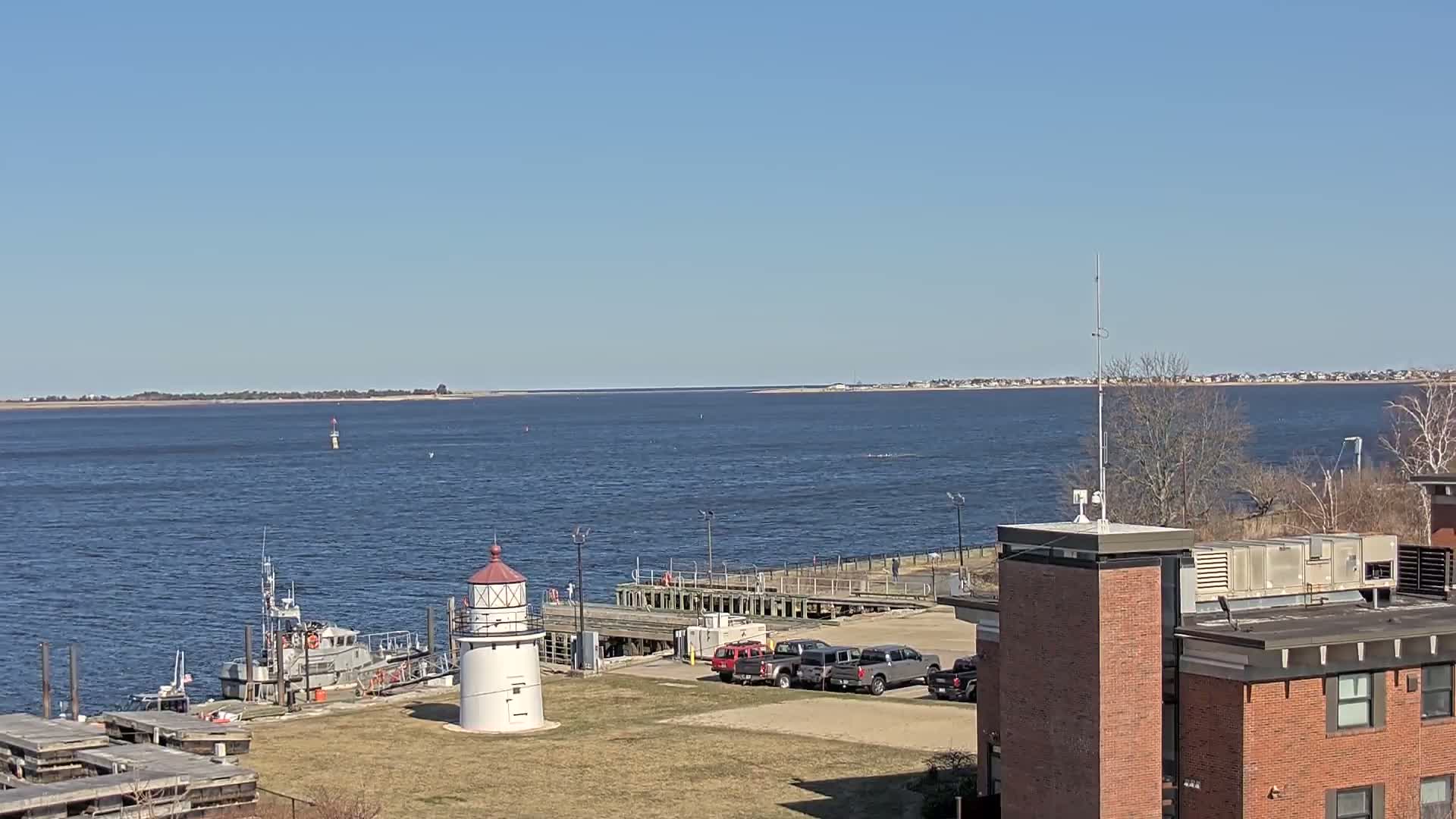 A small lighthouse stands near a docked boat and several vehicles, overlooking a calm body of water under a clear blue sky.