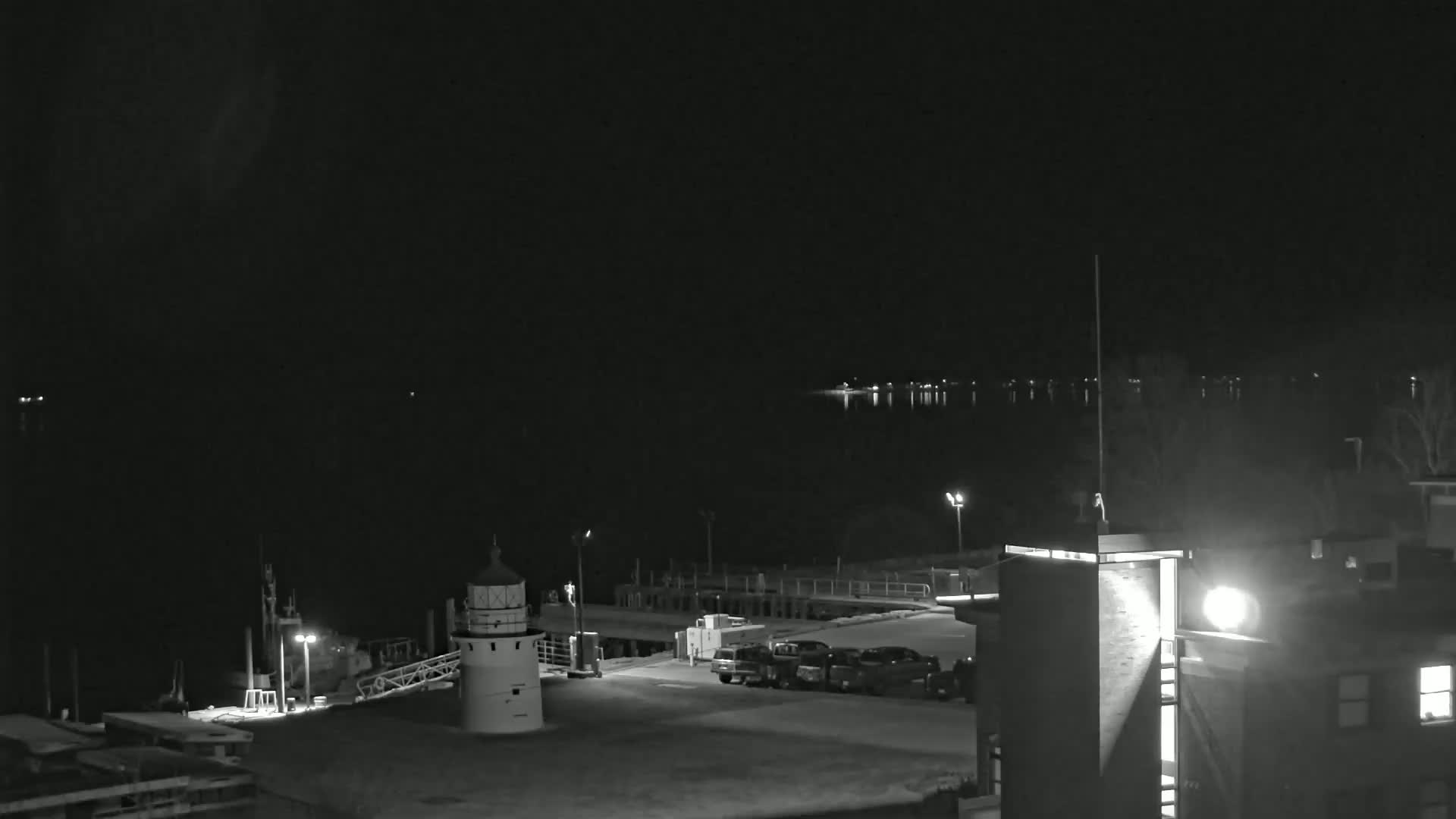 A nighttime, black and white view of a harbor with a lighthouse, several vehicles in a parking lot, and calm water reflecting lights across from the dock.