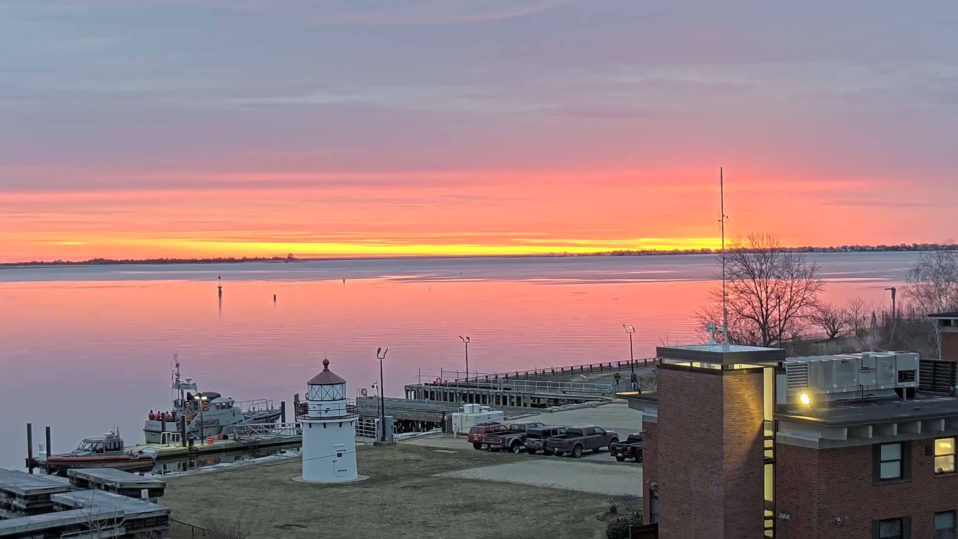 A pink and orange sunrise over calm water, reflecting the colors, with a small lighthouse, docked boats, and a building visible in the foreground.
