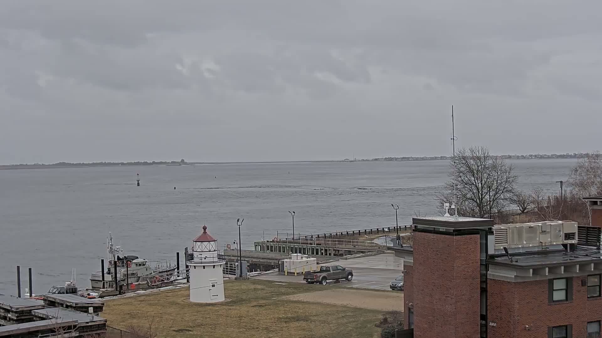 A small white lighthouse stands near a harbor with a docked boat, under an overcast sky, overlooking a wide body of water.
