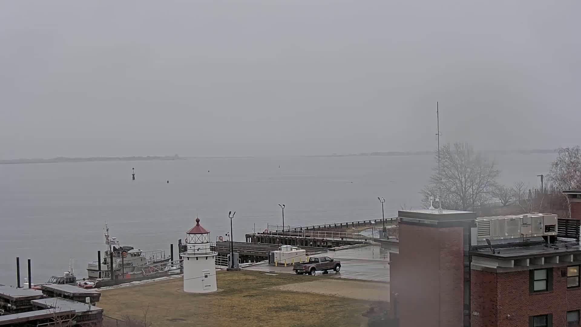 A small lighthouse and a docked boat are visible near a brick building on an overcast day beside a calm body of water.