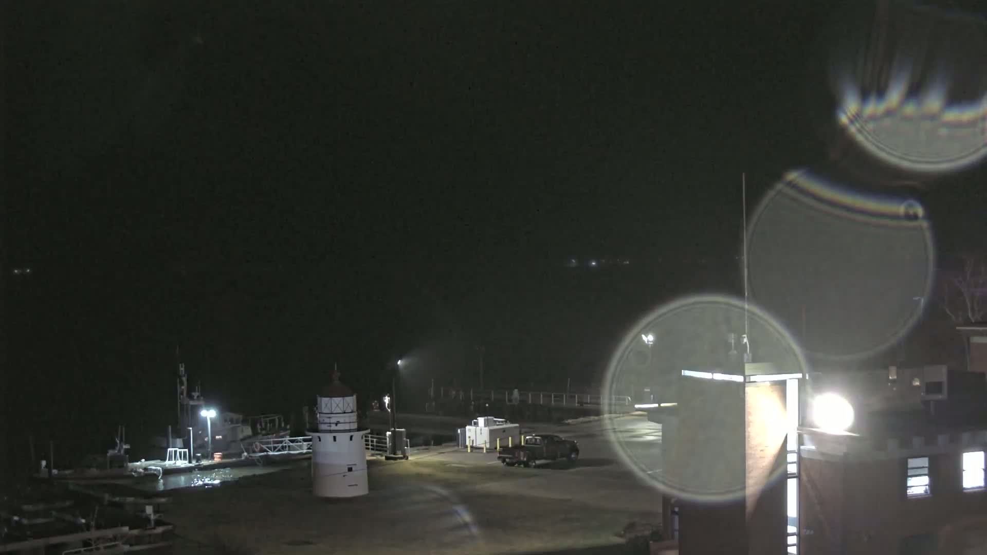 A nighttime view of a harbor with a lighthouse, boats, and a truck parked near a building, under a dark, possibly cloudy sky.