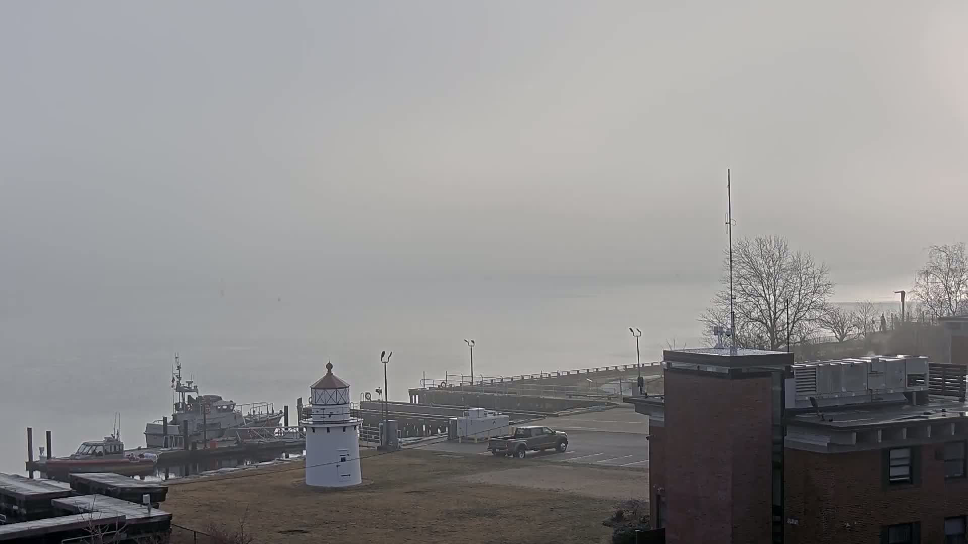 A fog-shrouded harbor scene features a lighthouse, docked boats, a truck in a parking lot, and a brick building with equipment on its roof.
