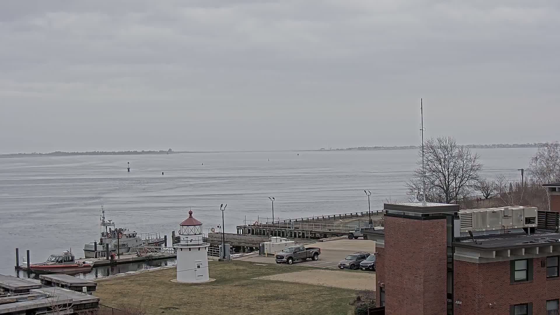 A lighthouse and Coast Guard boat are docked near a parking lot and brick buildings overlooking a calm, gray, overcast body of water.