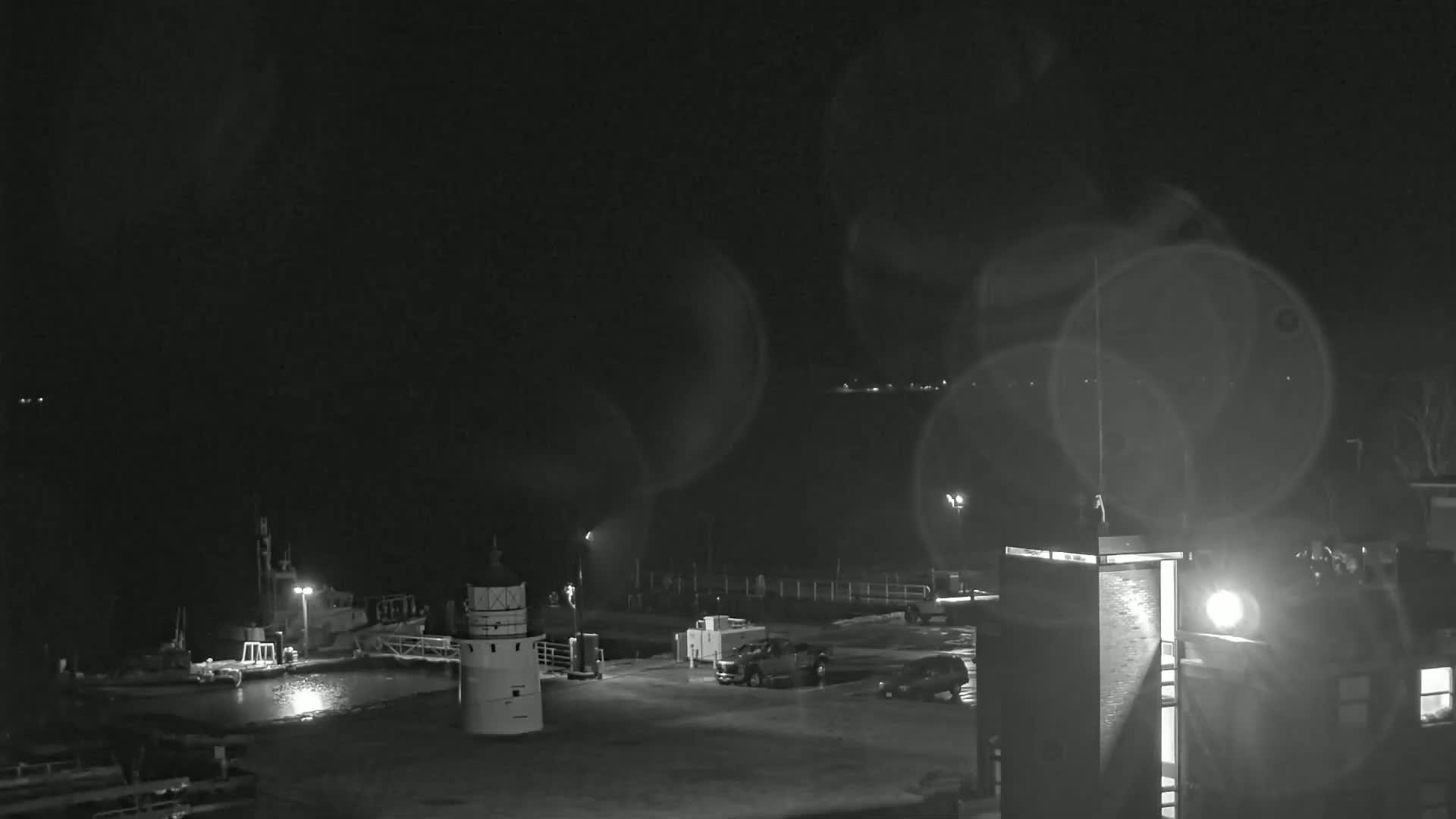 A nighttime, slightly hazy view of a harbor with boats docked, a small lighthouse, and vehicles parked in a lot adjacent to a tall building.