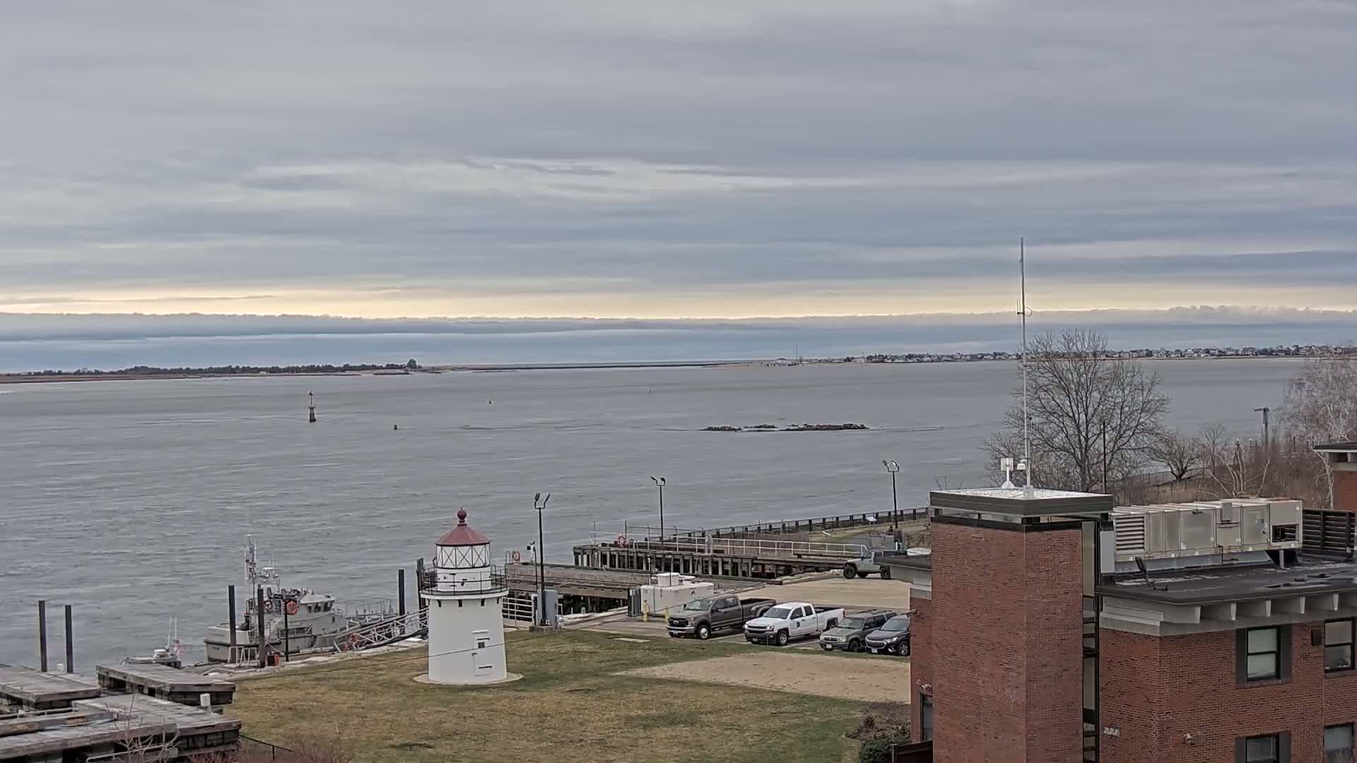 A small lighthouse sits on a grassy area overlooking a calm, gray body of water under a cloudy sky; a small boat is moored nearby, and several vehicles are parked near a brick building in the foreground.