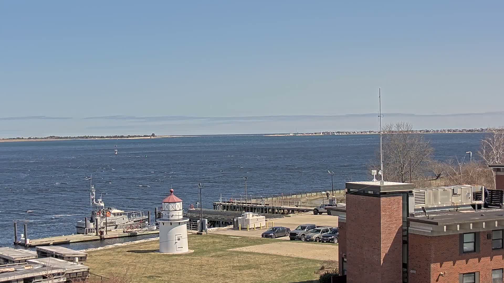 A small lighthouse and a docked boat are situated near a brick building overlooking a body of water on a partly sunny day with a light wind.