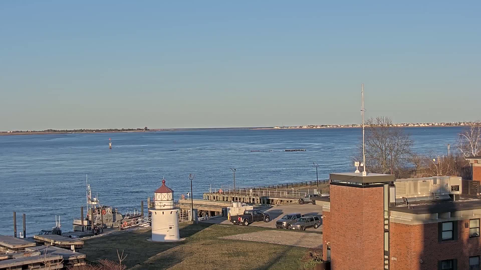A small white lighthouse stands near a dock with several vehicles parked nearby, overlooking a calm body of water under a clear blue sky.