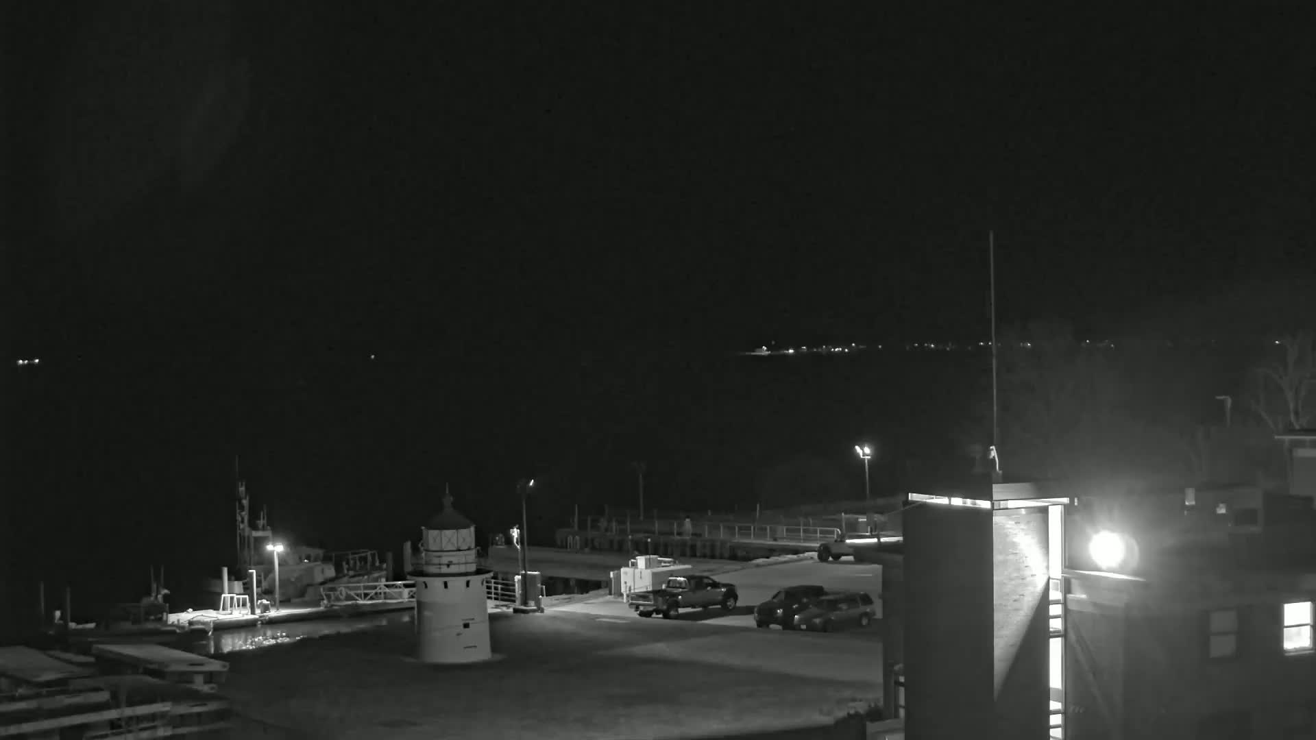 A nighttime, black-and-white view of a lighthouse and harbor with a few vehicles parked nearby.