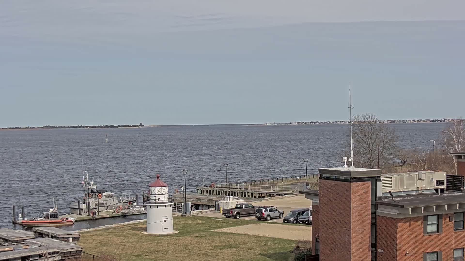 A small lighthouse and Coast Guard vessels are docked at a waterfront facility on a calm, overcast day, with a wide expanse of water and distant shoreline visible in the background.