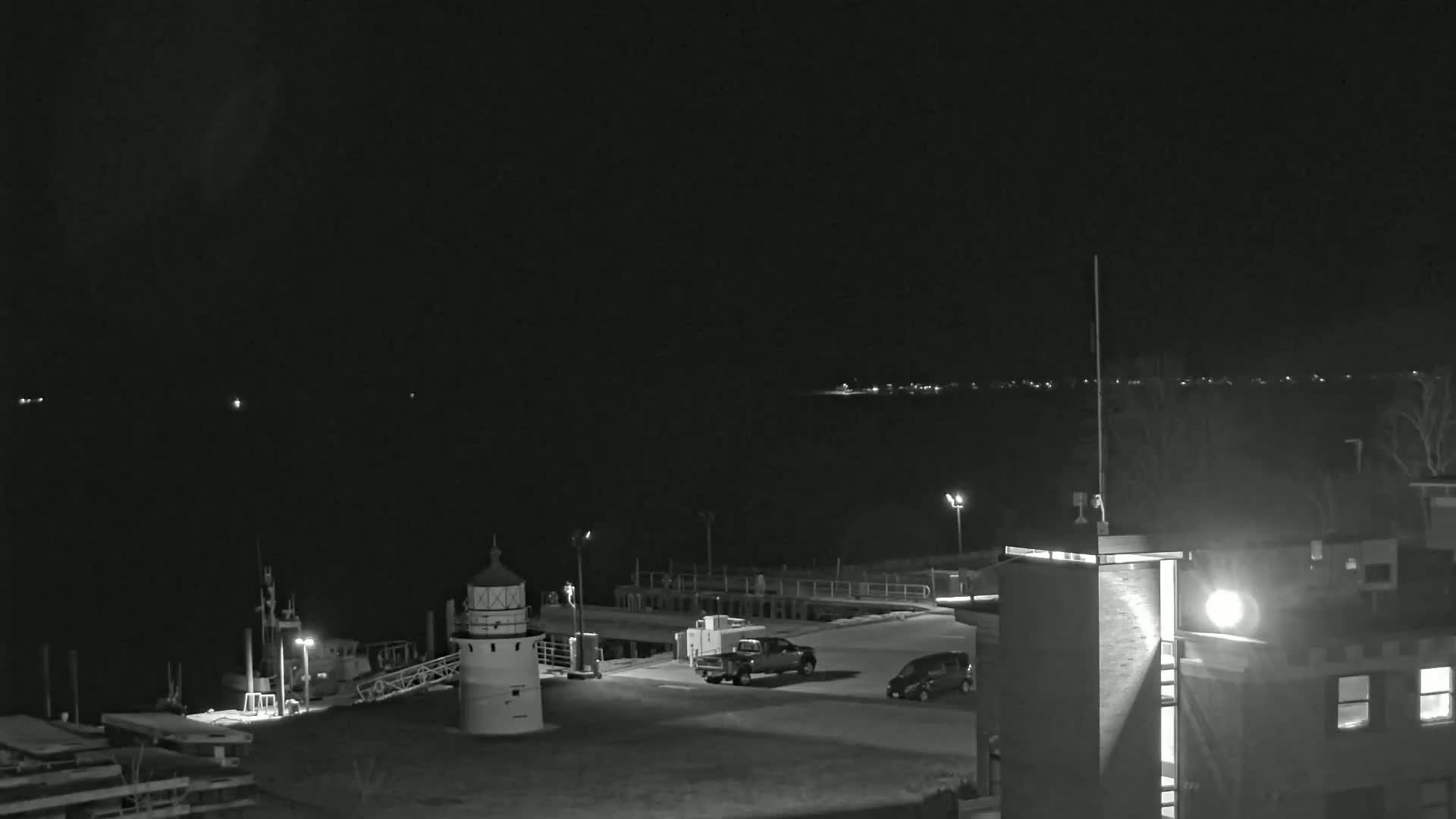 A nighttime, black and white view of a harbor with a lighthouse, a small boat, and vehicles parked in a lot, under a dark, clear sky.