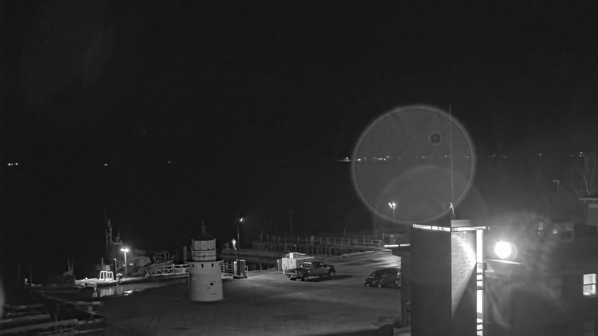 A nighttime, black-and-white view of a harbor with a lighthouse, boats, and vehicles parked in a lot near a building.