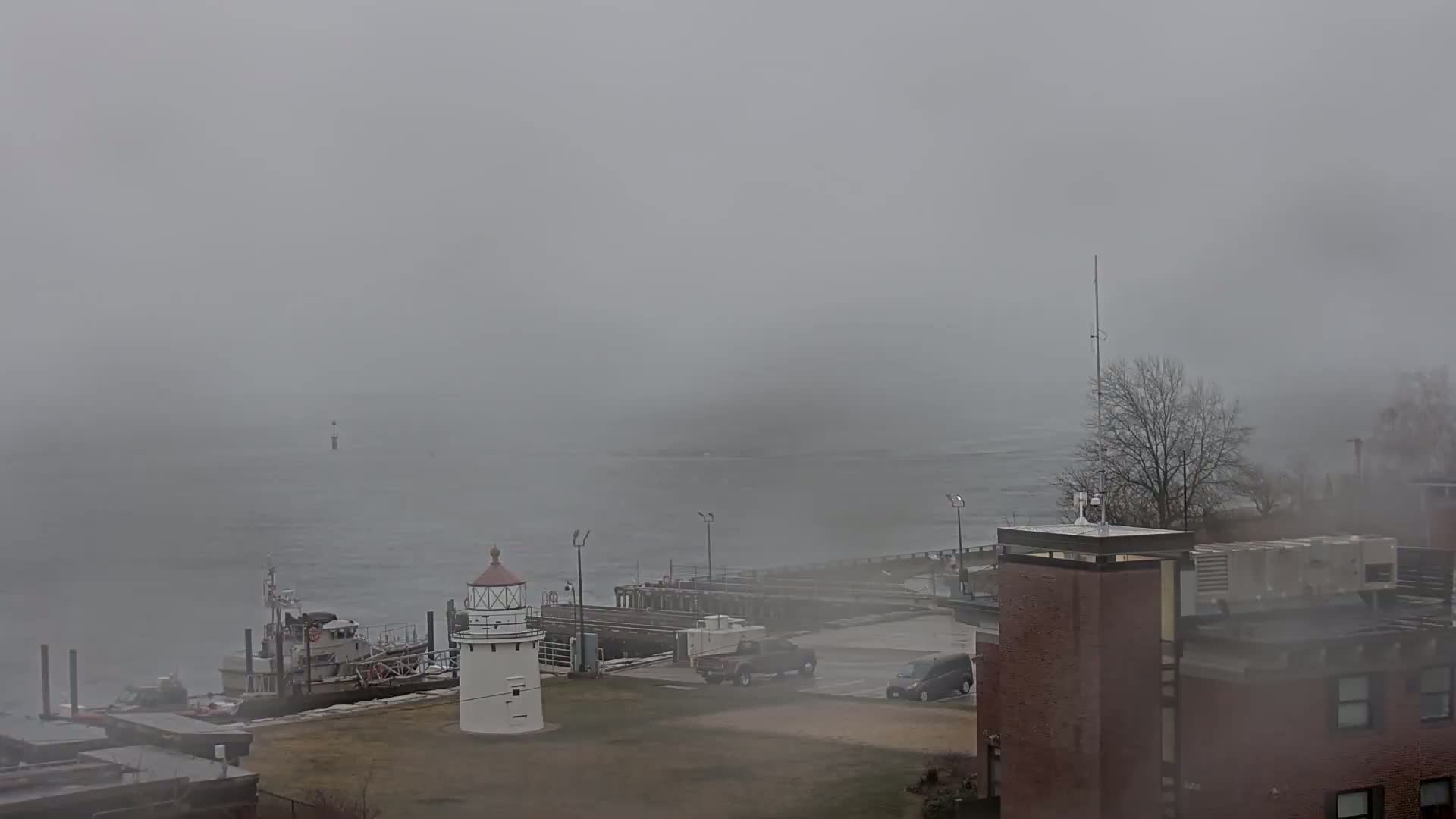 A small lighthouse sits on a foggy waterfront next to a docked boat and a building, with a few vehicles visible in the parking lot.