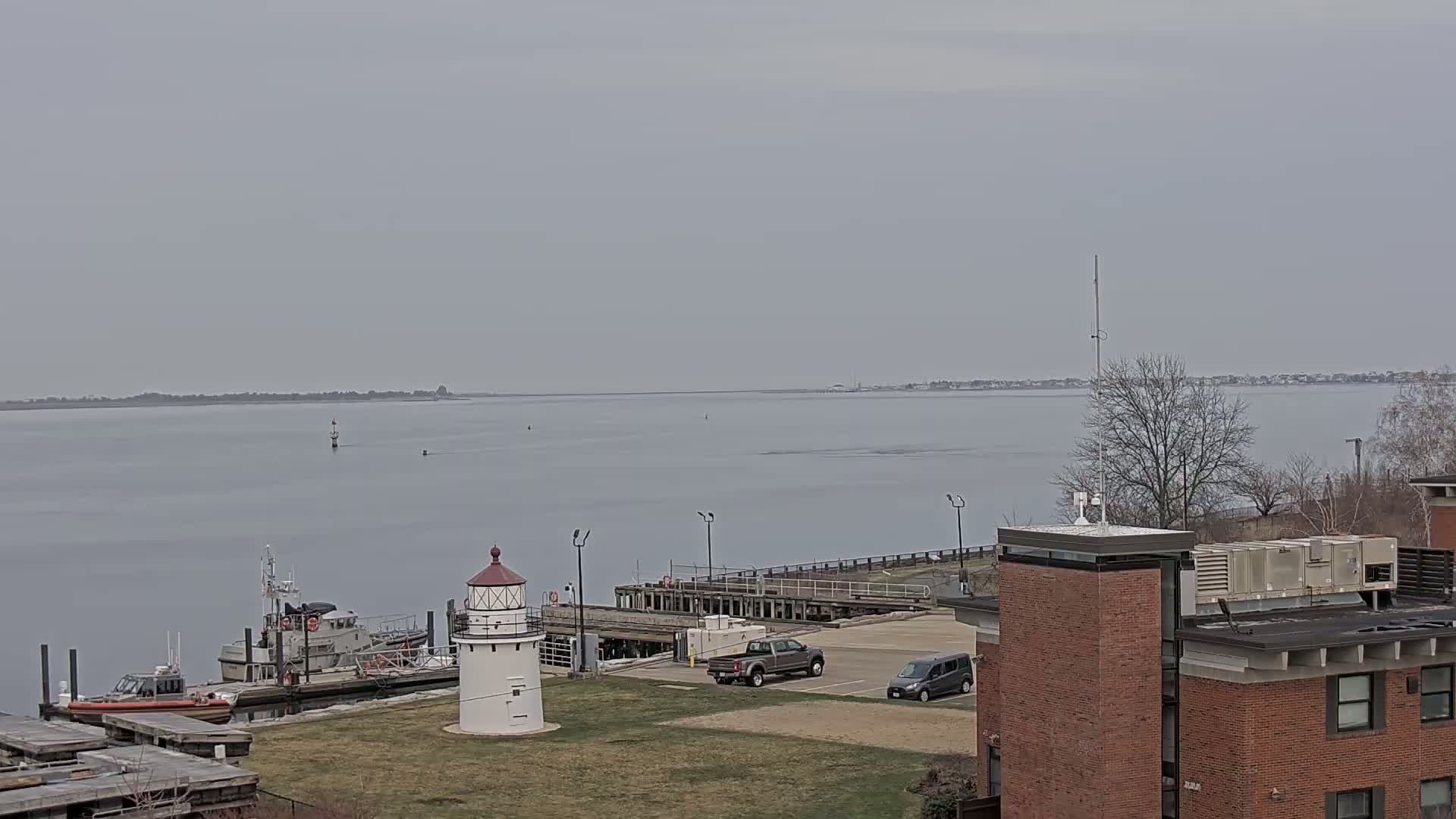 A small lighthouse sits on a grassy area near a dock with several boats and vehicles, overlooking a calm, gray body of water on an overcast day.
