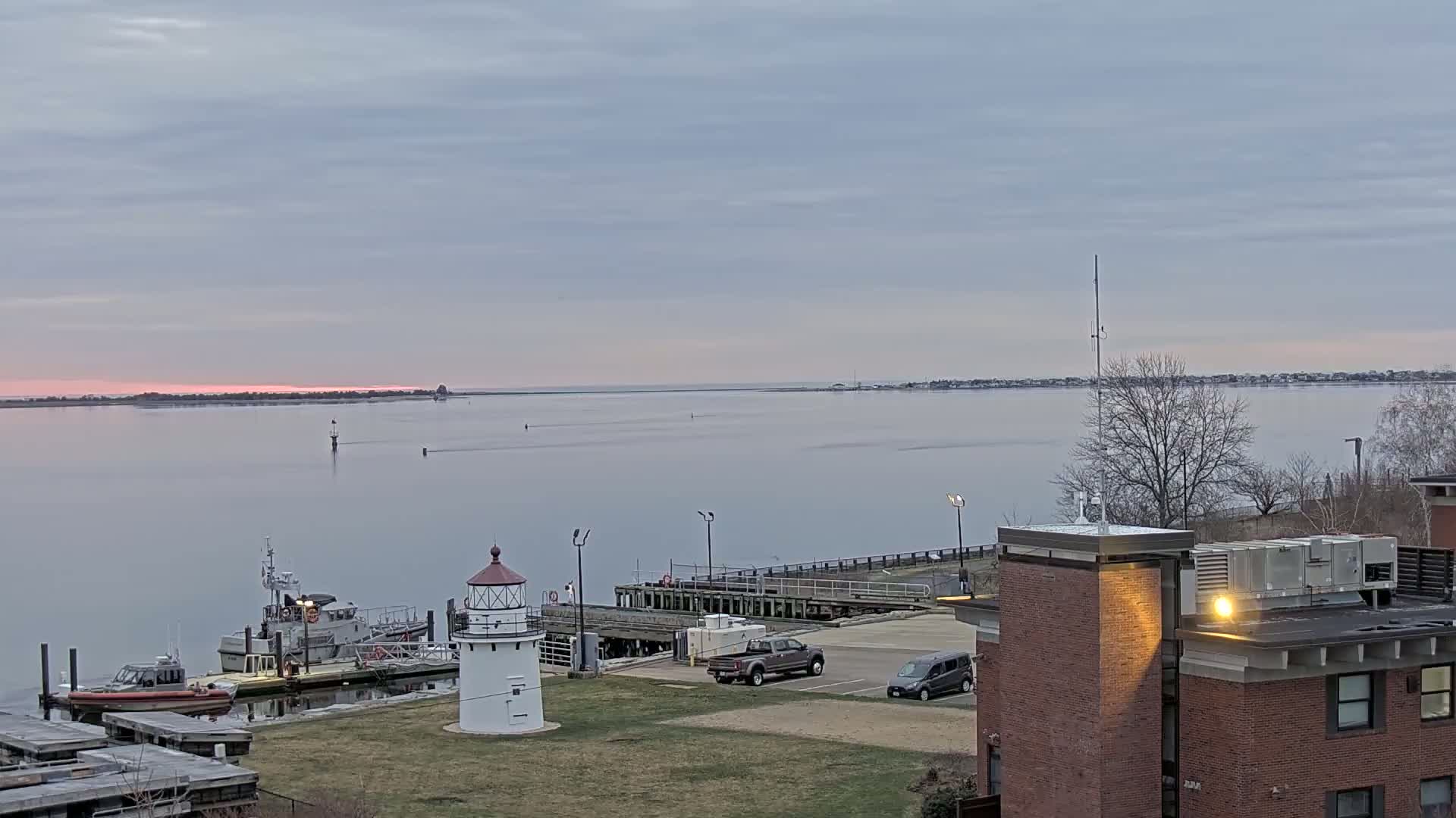 A calm body of water at sunset is viewed from a high vantage point, showing a lighthouse, small boats docked at a pier, a few vehicles in a parking lot, and a brick building in the foreground.