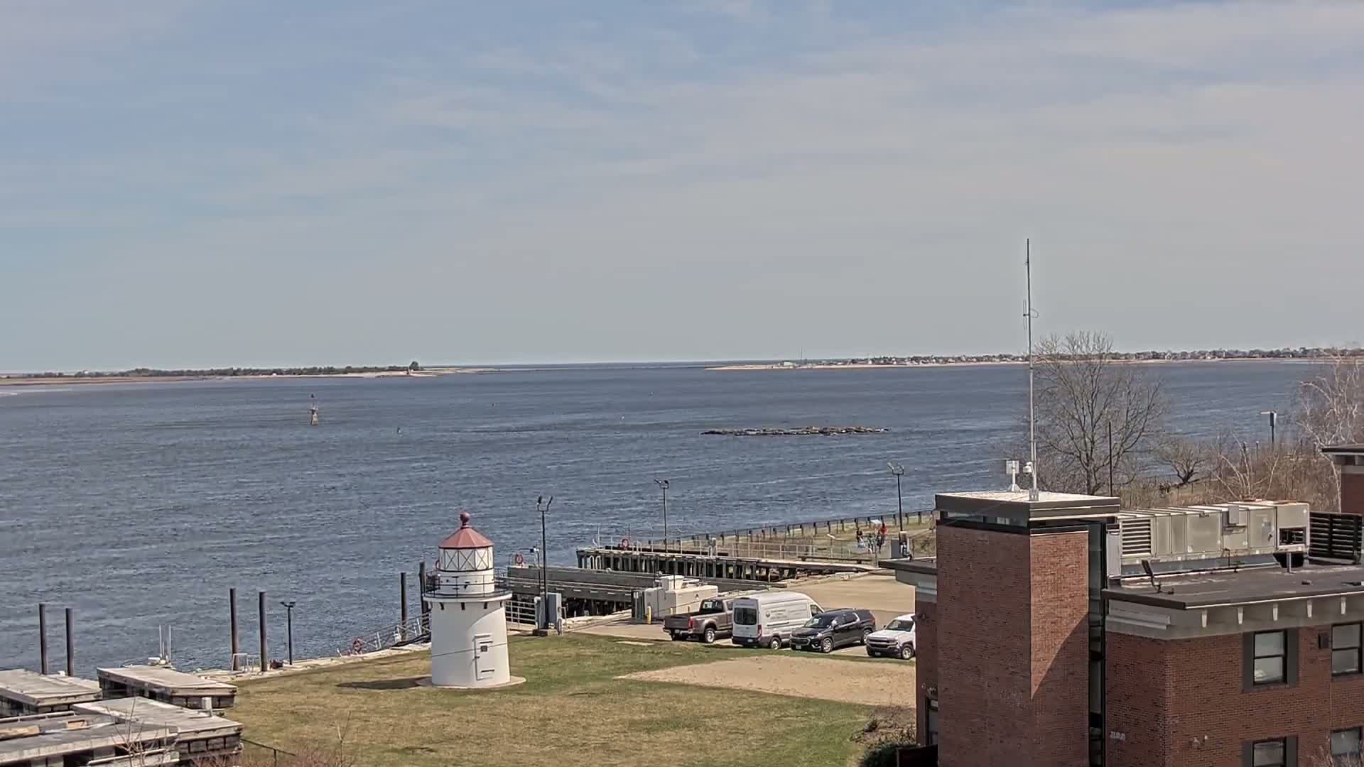 A small lighthouse stands on a grassy area overlooking a calm body of water under a mostly sunny sky, with a few parked vehicles and a larger brick building visible in the foreground.