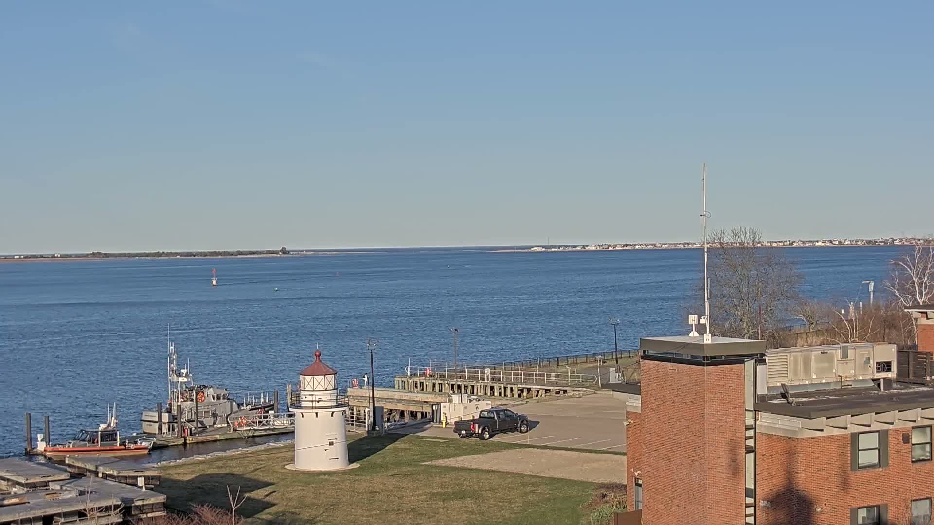 A calm body of water stretches to a distant shoreline under a clear blue sky, with a small lighthouse, docked boats, and a brick building in the foreground.