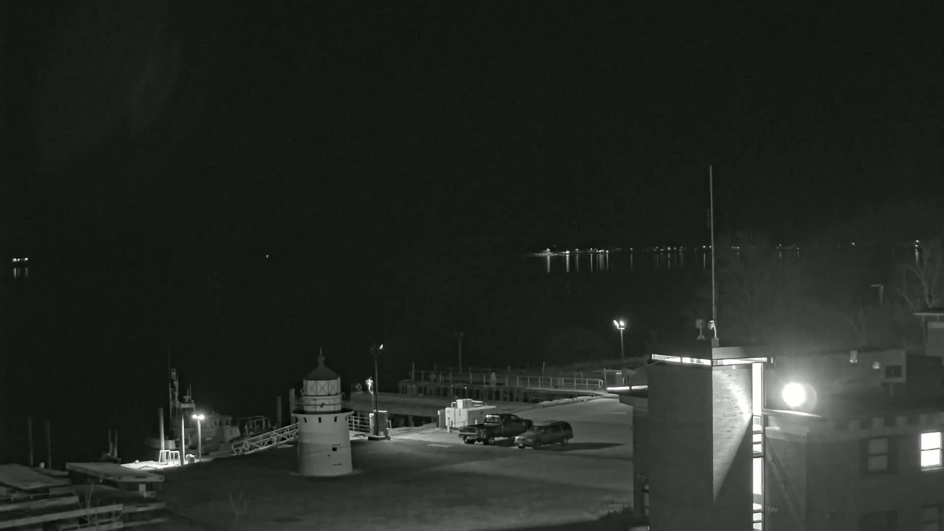 A nighttime, black and white view of a lighthouse, a small dock, two vehicles in a parking lot, and a building next to a calm body of water under a dark sky.