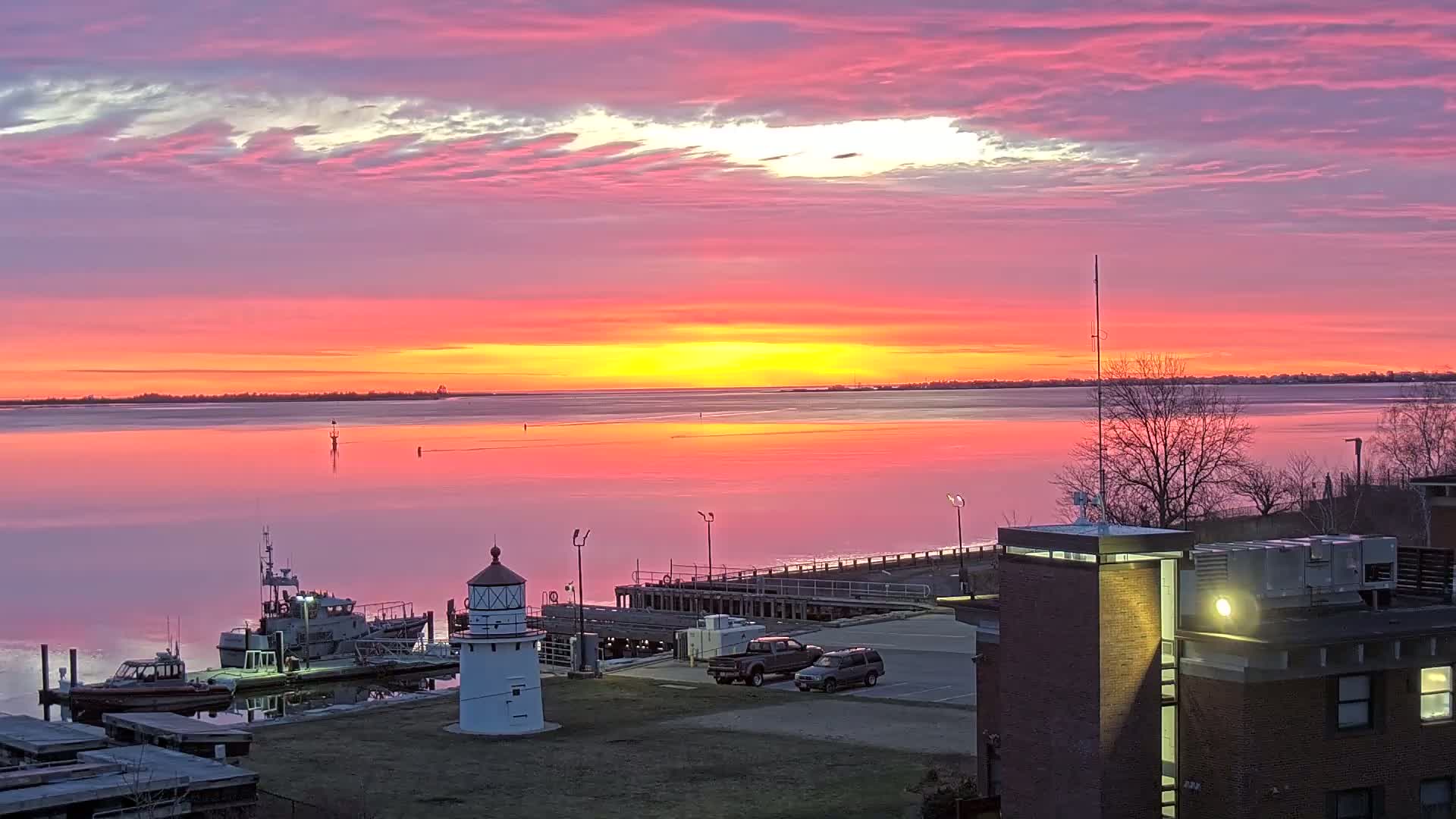 A vibrant pink and orange sunrise reflects on calm water, illuminating a small lighthouse, docked boats, and a parking lot with vehicles near a brick building.