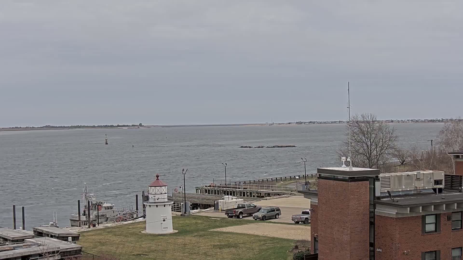 A small lighthouse and a few parked vehicles are situated near a dock on a body of water under an overcast sky.