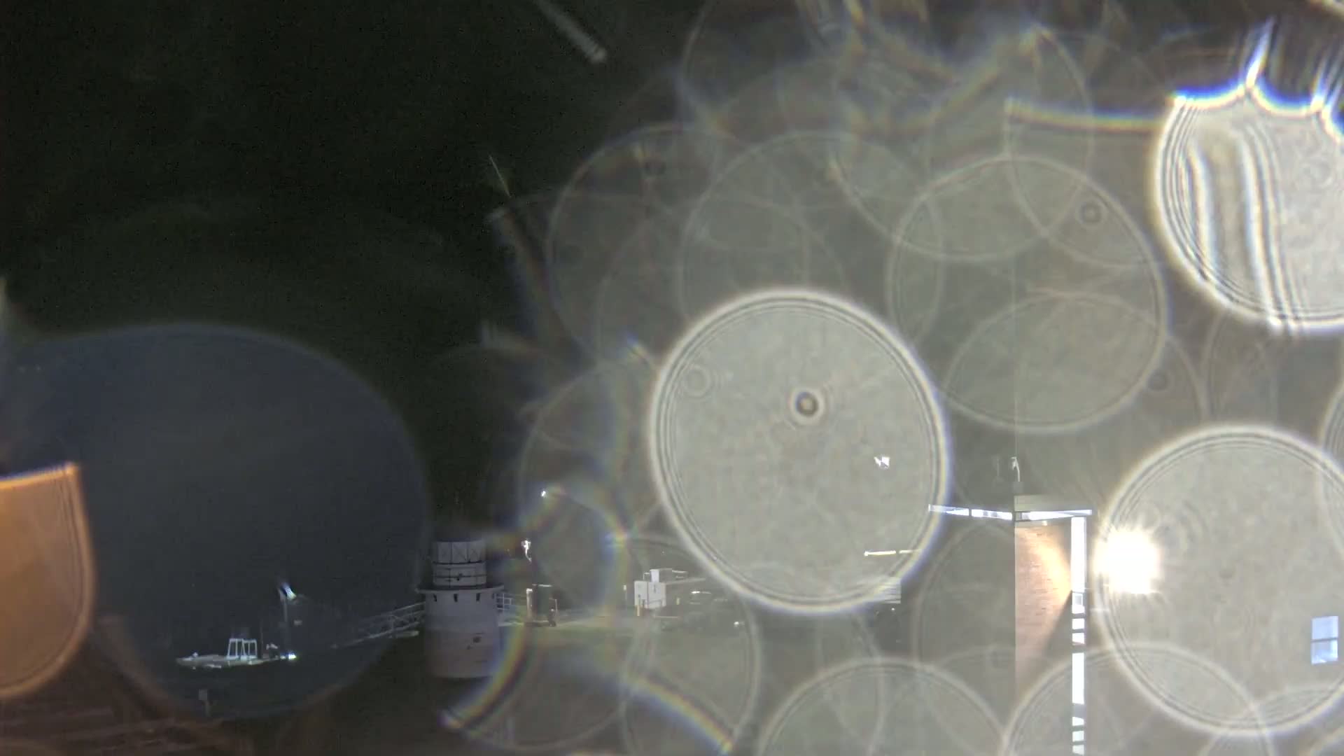 A nighttime view of a building and vehicles is seen through a blurry, distorted lens, showing numerous overlapping circular shapes.