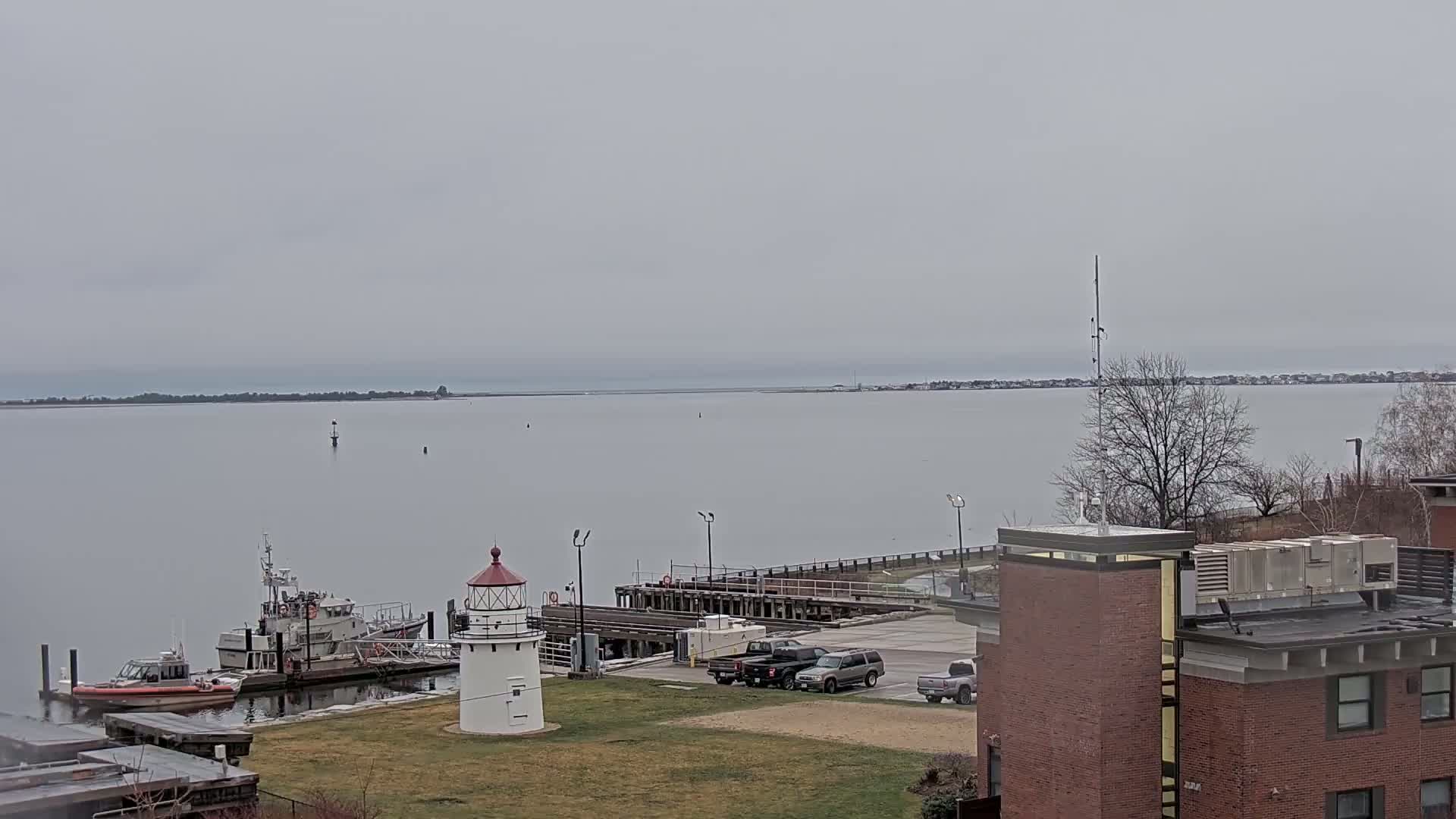 A small lighthouse stands on a grassy area next to a harbor with several boats and cars parked nearby, all under an overcast sky.