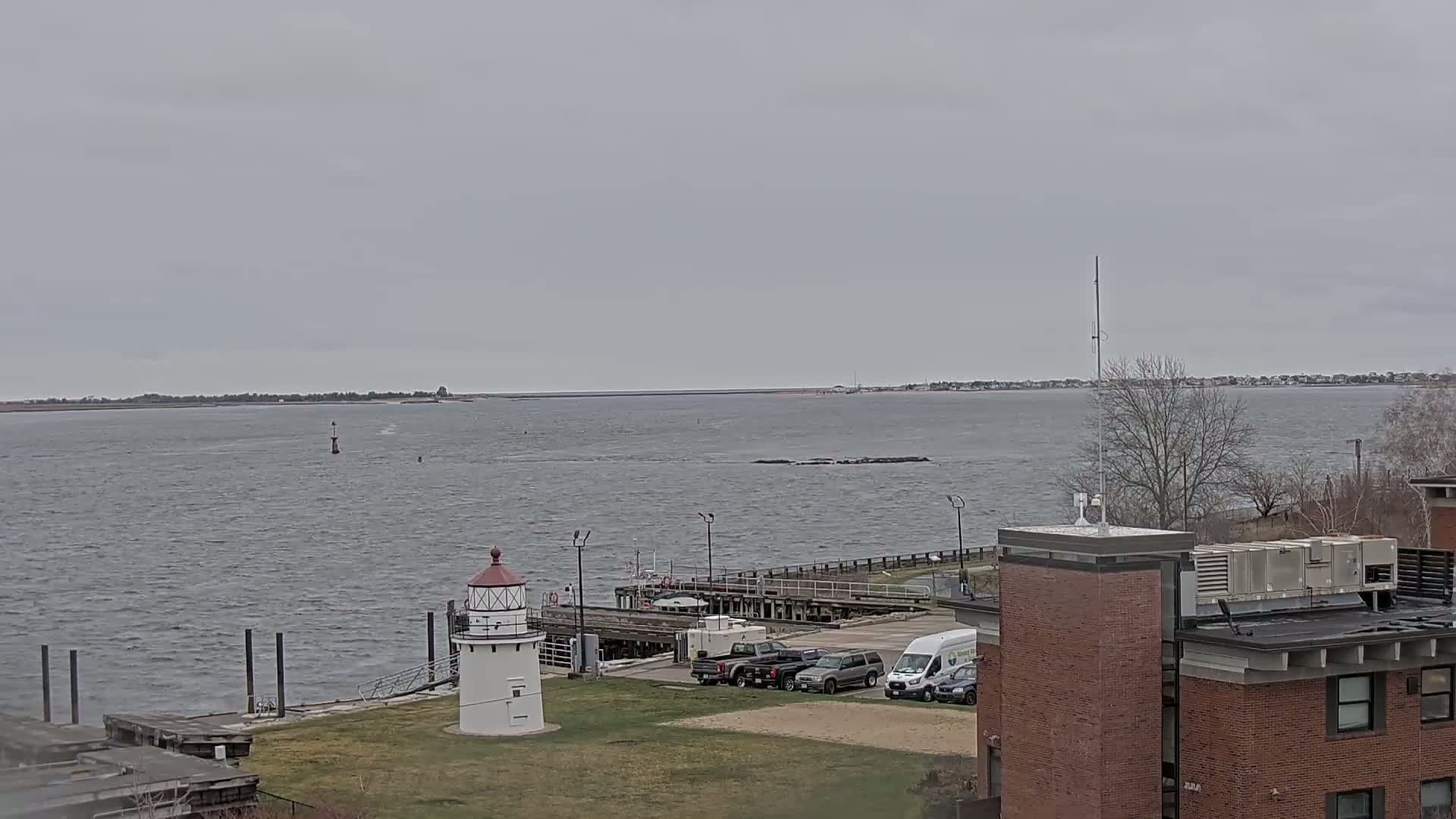 A small lighthouse sits on a grassy area next to a dock with several vehicles parked nearby, overlooking a choppy body of water under an overcast sky.