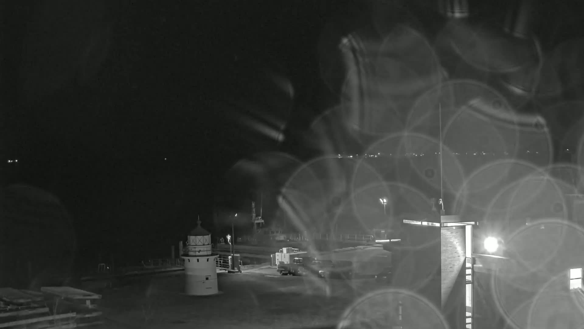 A nighttime view of a harbor with a lighthouse, vehicles, and buildings, seen through a rain-streaked window.