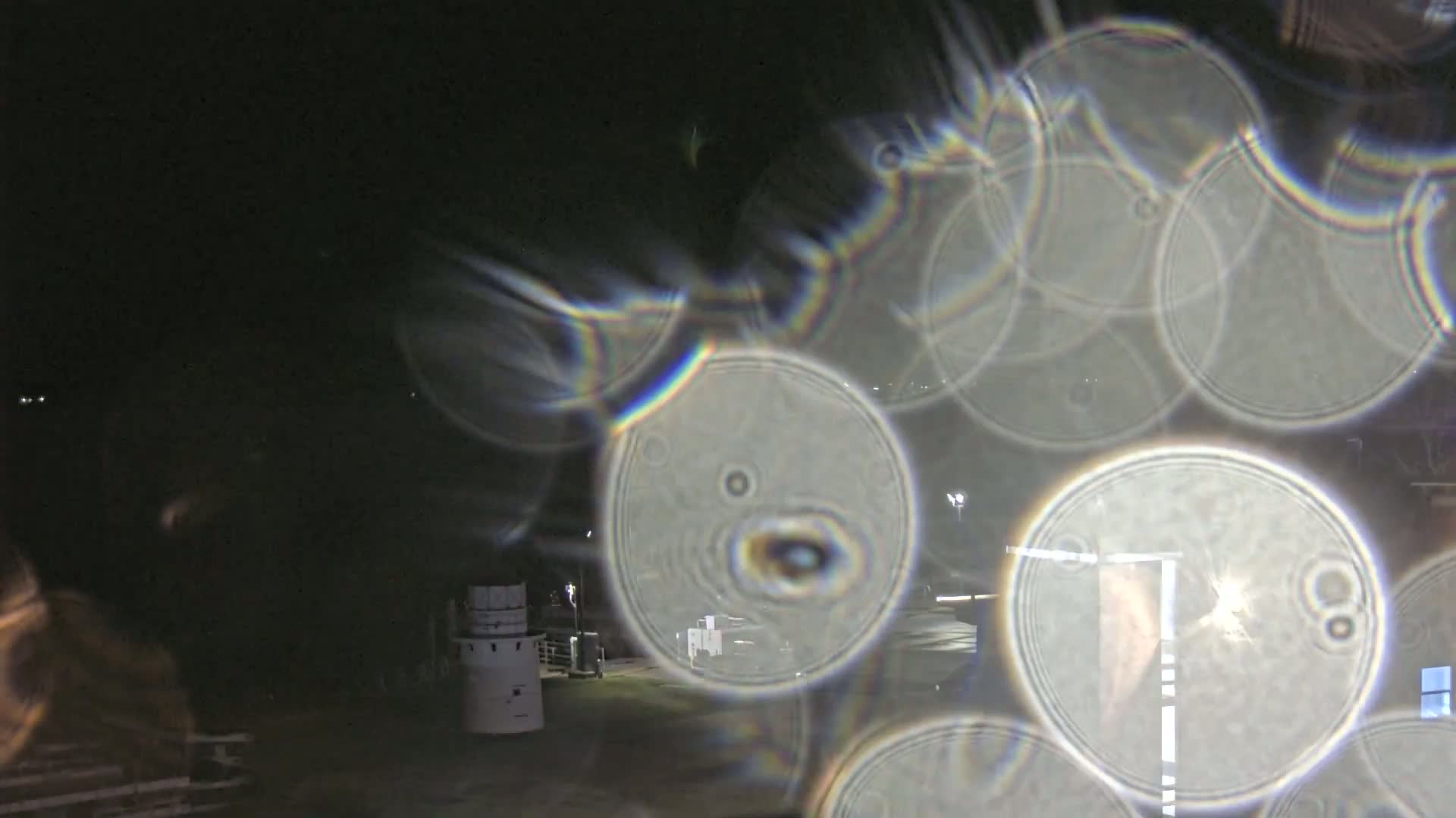 A nighttime outdoor scene is viewed through a surface that distorts the image into numerous overlapping, iridescent, circular shapes, obscuring the view of a building and roadway.