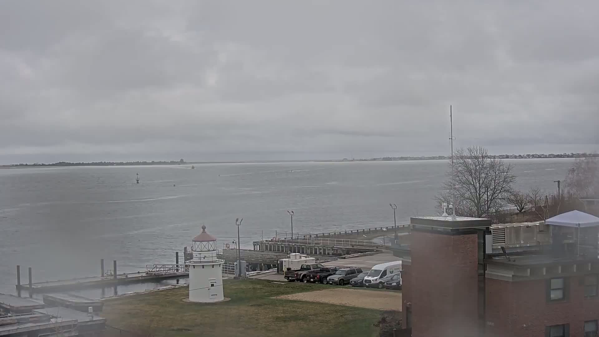 A white lighthouse stands on a grassy area next to a dock and body of water under an overcast sky.