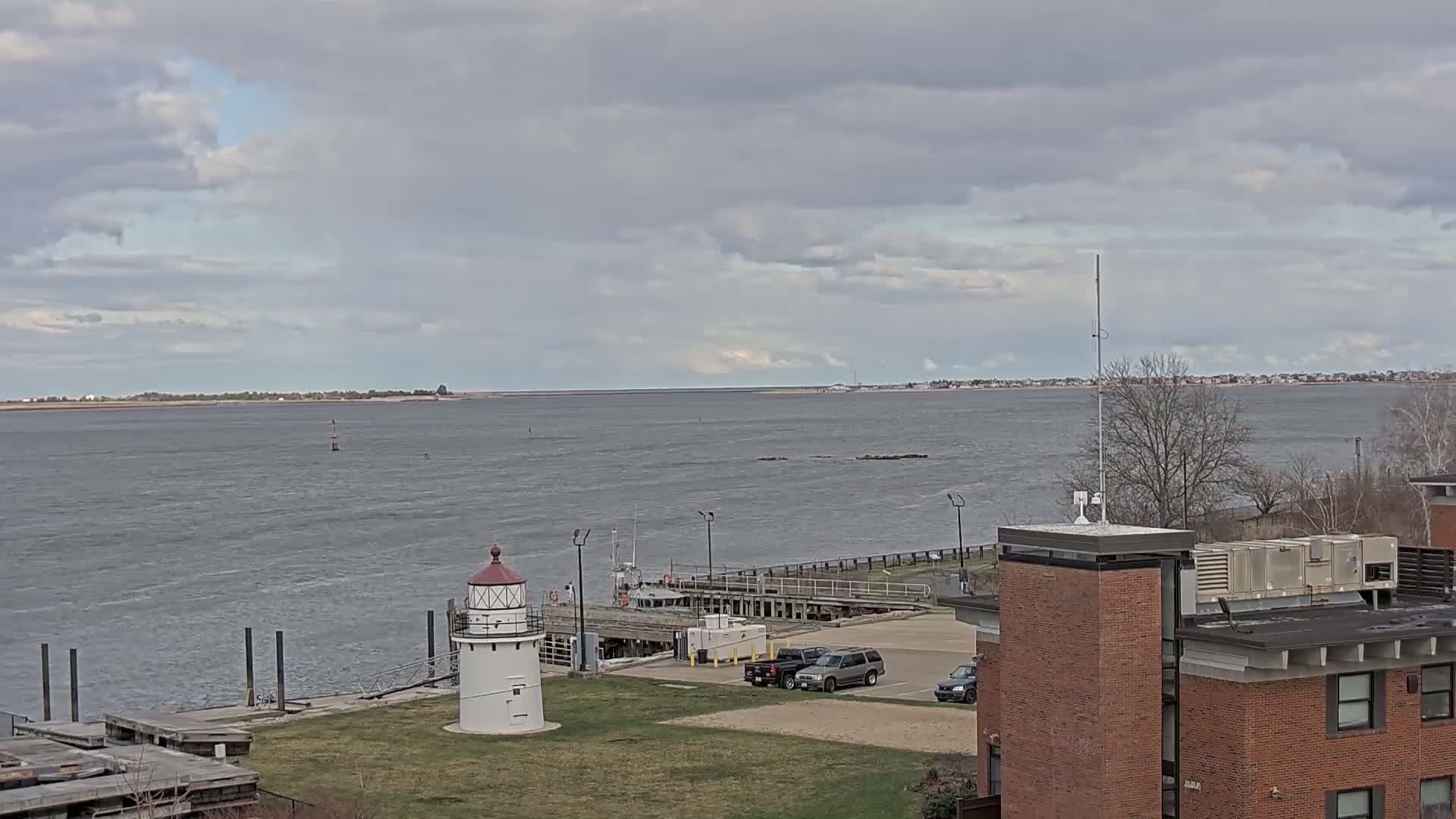 A small lighthouse stands near a dock and parking lot overlooking a body of water under a cloudy sky.